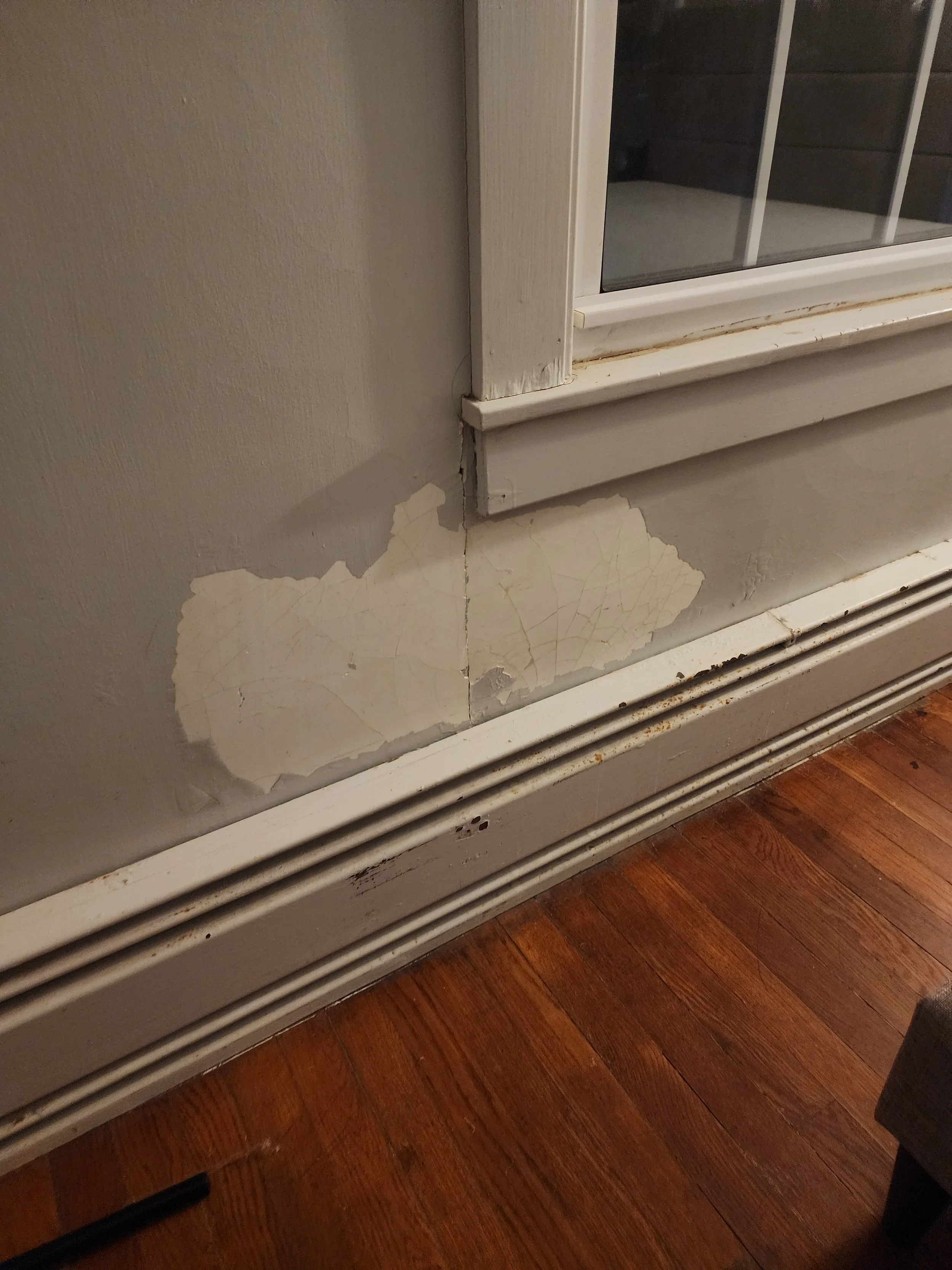 Peeling paint and damage on the lower part of a wall near a baseboard heater and window. The wall is beige, and the hardwood floor is reddish-brown.