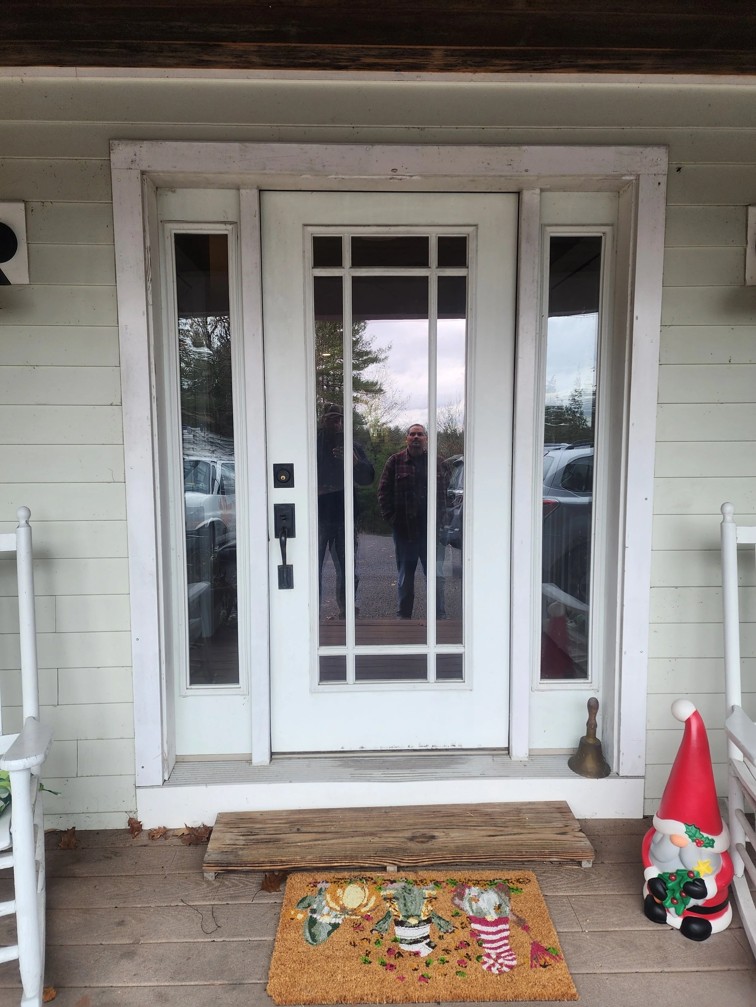 A glass door with sidelights and decorative glass panels, reflecting trees and parked cars. There are two men visible in the reflection, one wearing a plaid shirt and the other in a jacket. The door has a doorbell and handle, with a small bell on the
