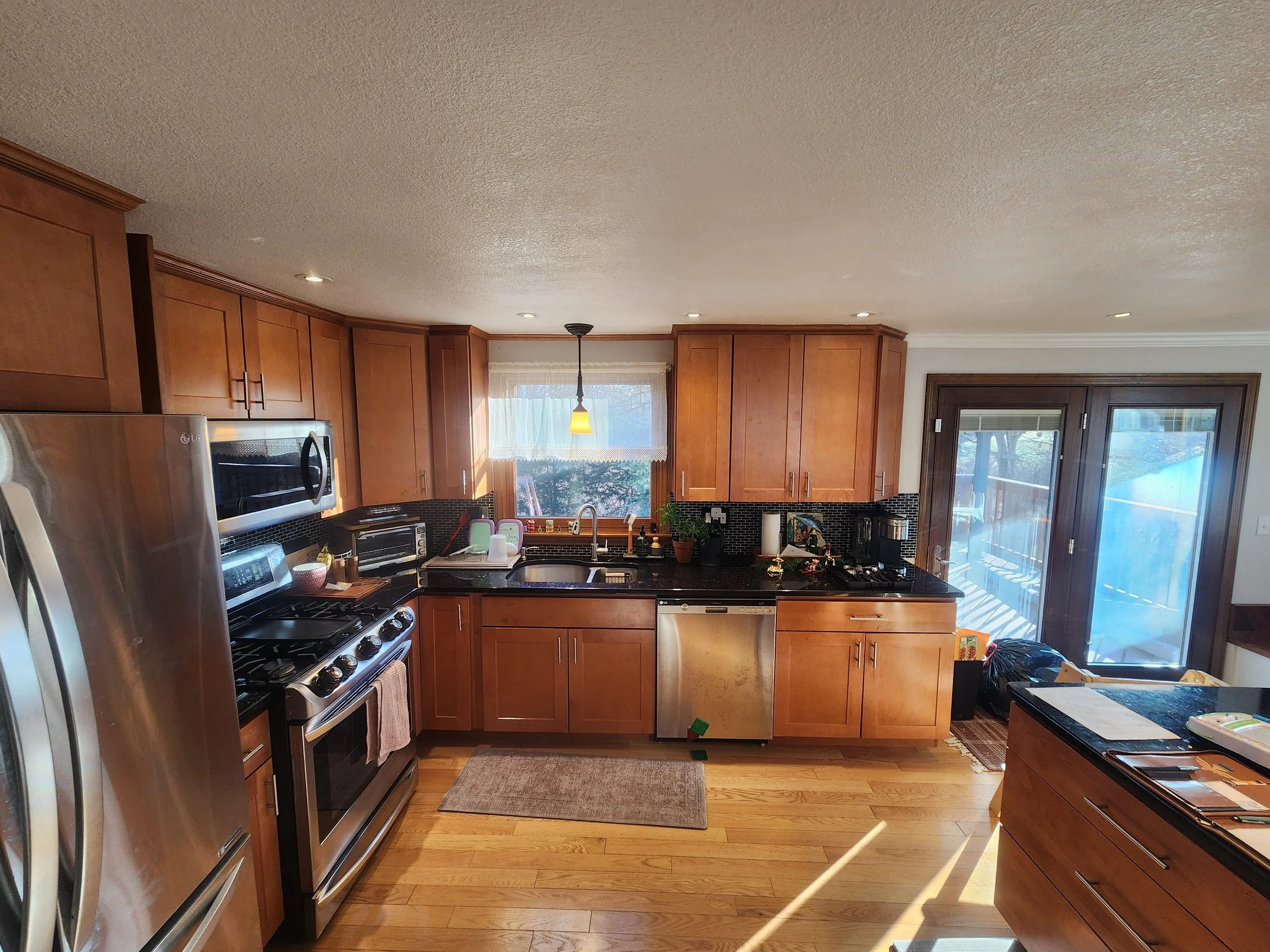 Kitchen with wooden cabinets, stainless steel appliances, black countertops, natural light from window and sliding glass door, hardwood floor, small rug, various appliances and items on counters.