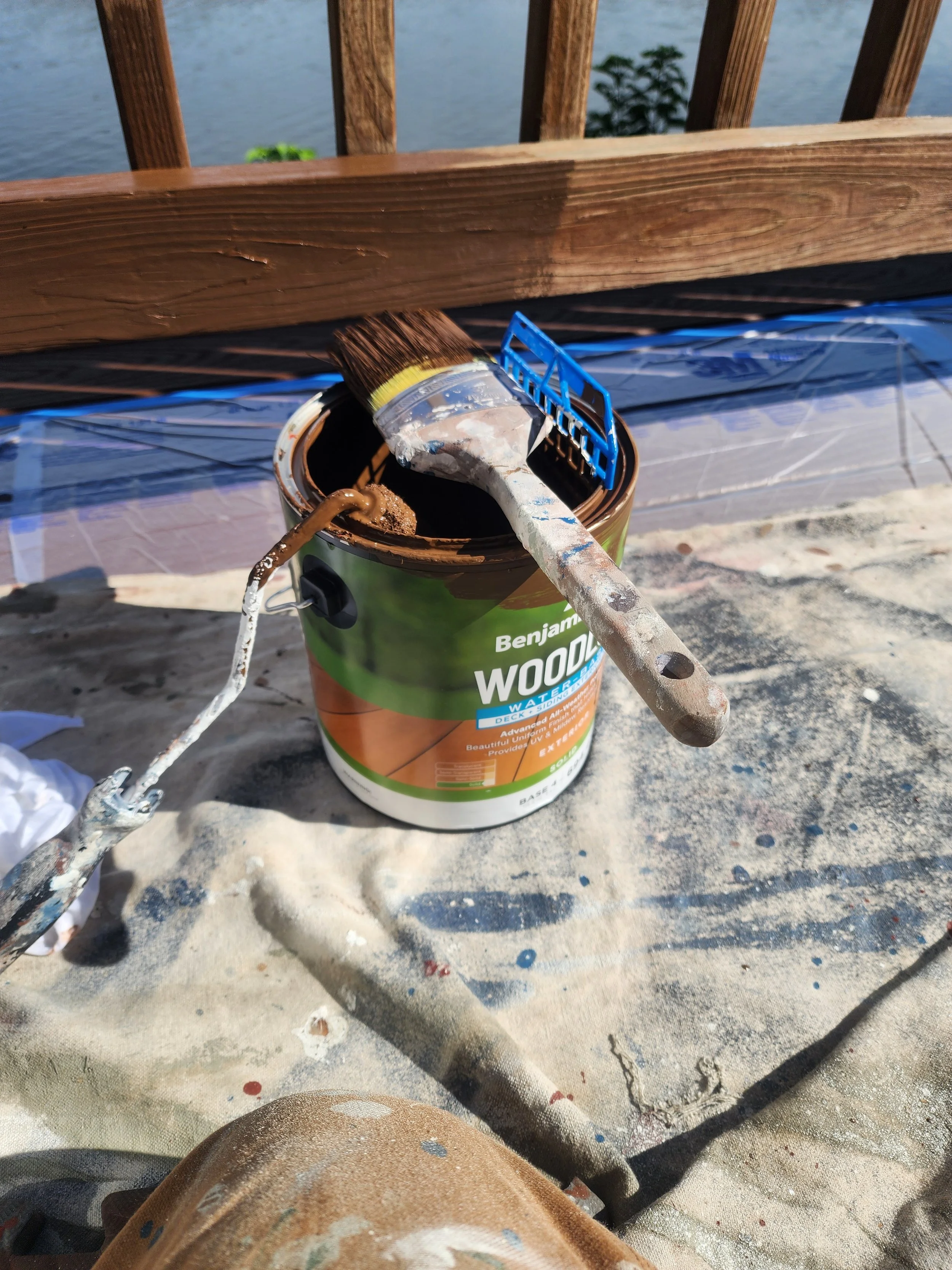 Paintbrush and paint roller resting on a can of wood stain, with a sanded surface and paint splatters around. In the background, a wooden railing and a body of water are visible.