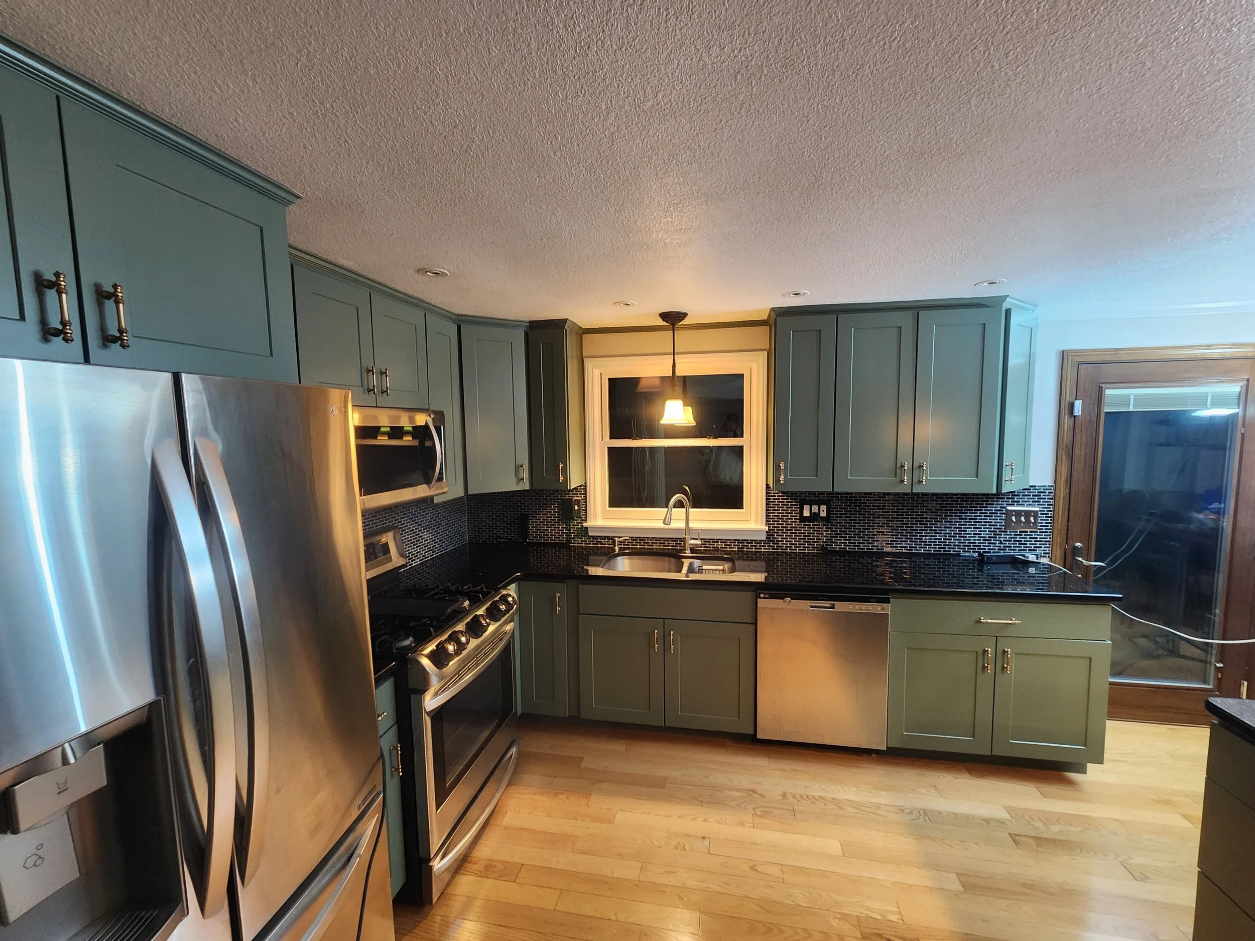 A kitchen with green cabinets, black countertops, and stainless steel appliances, including a refrigerator, stove, microwave, and dishwasher. There is a window above the sink and a sliding glass door to the right.