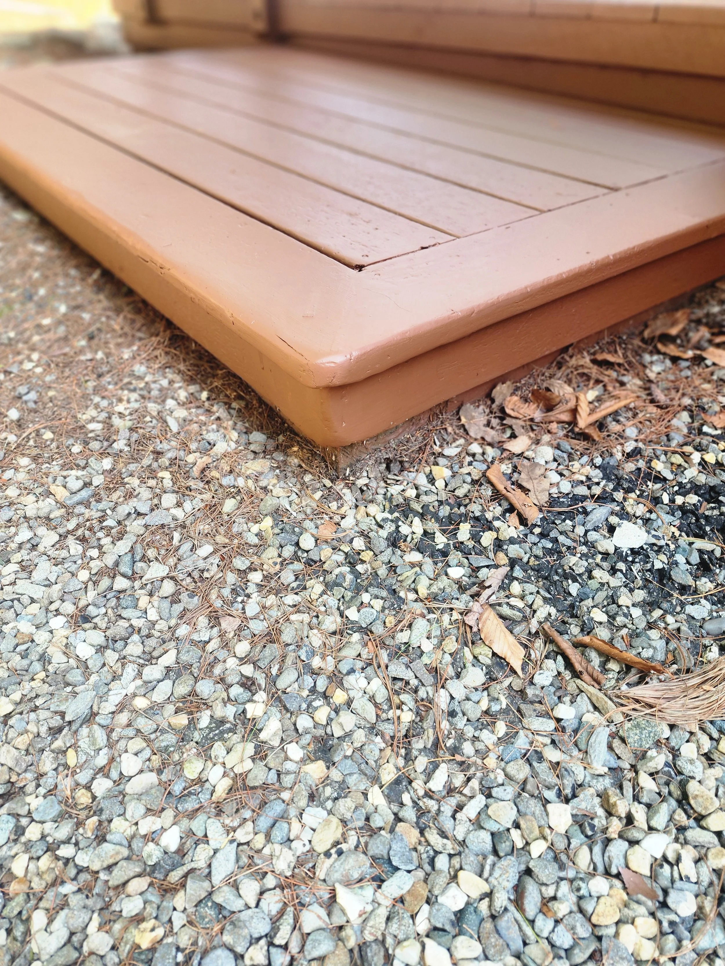 Close-up of a brown wooden deck step on gravel ground with small rocks and dried leaves.