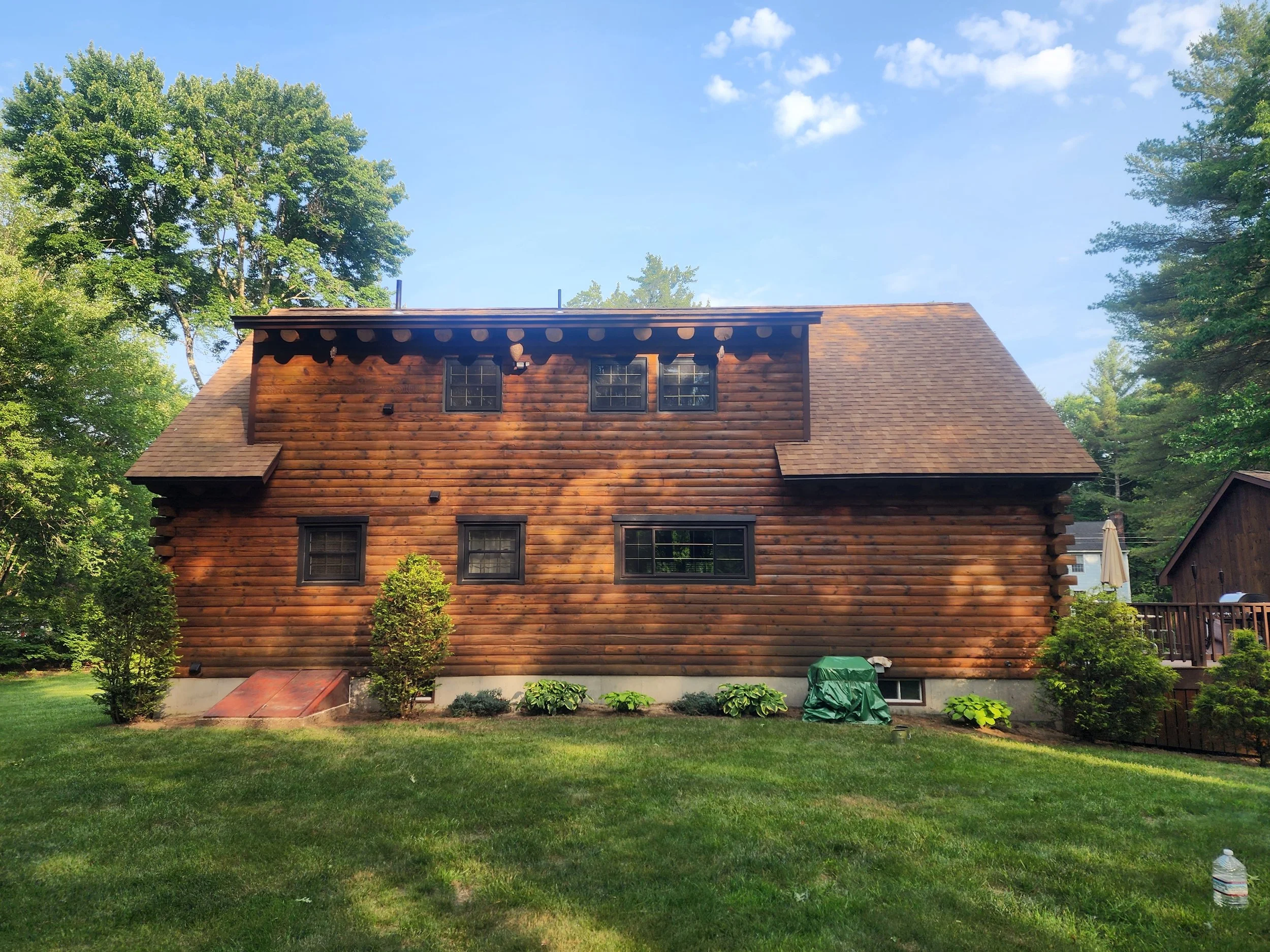 Backyard view of a two-story log cabin with small windows, surrounded by lush green trees and grass, under a partly cloudy sky.