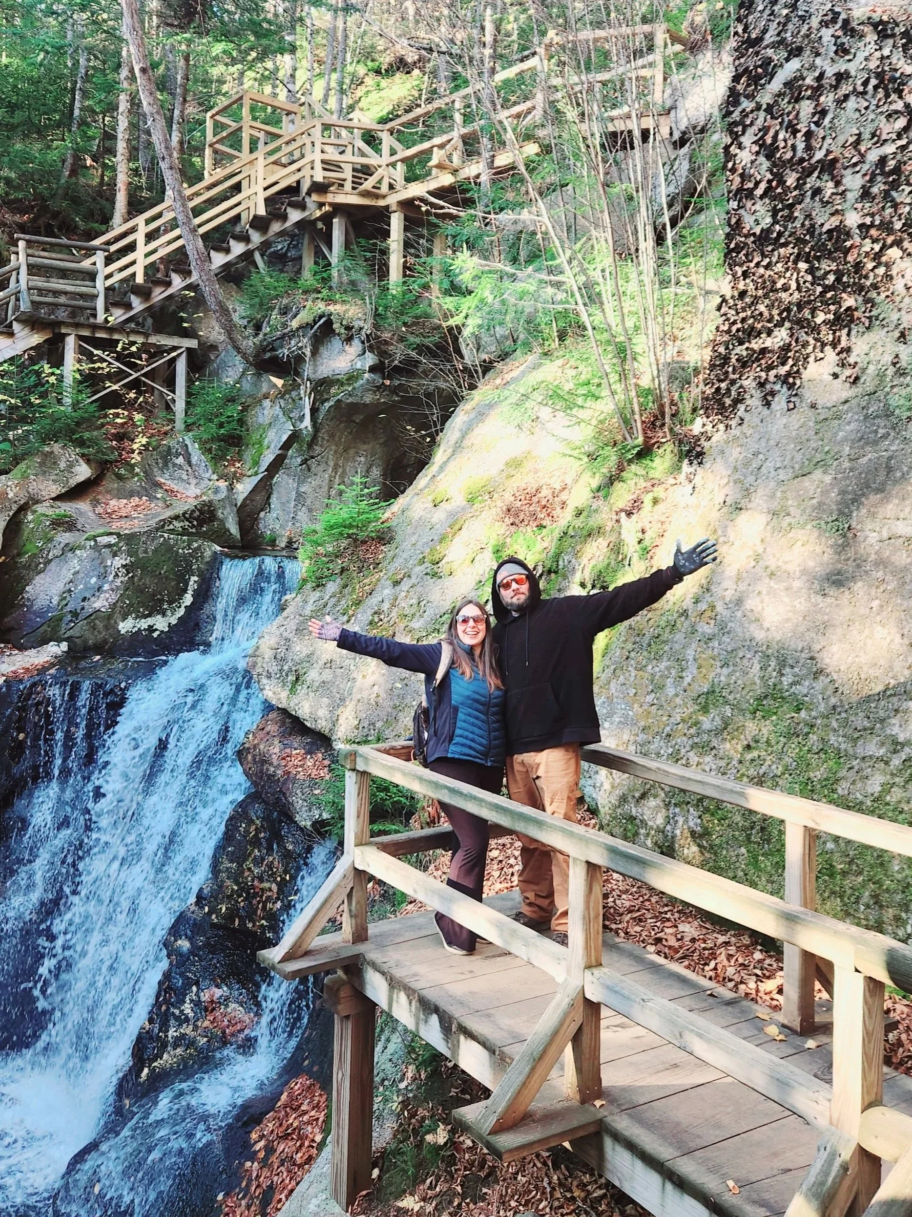 Two people standing on a wooden platform with arms open wide, in front of a waterfall and lush green forest.