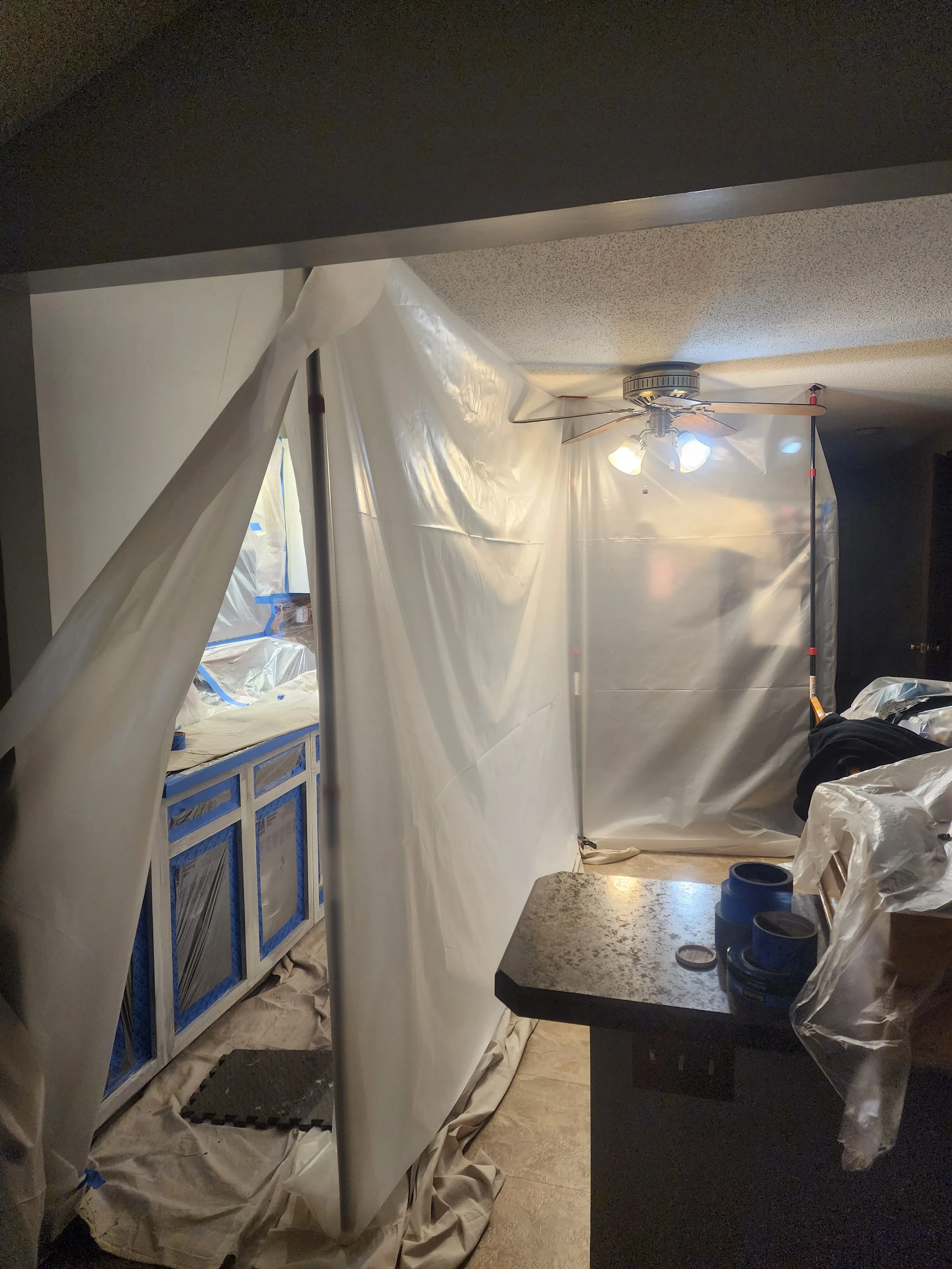 A room under renovation with plastic sheeting covering the walls and furniture, a ceiling fan with lights, and tools and supplies scattered on the countertop.