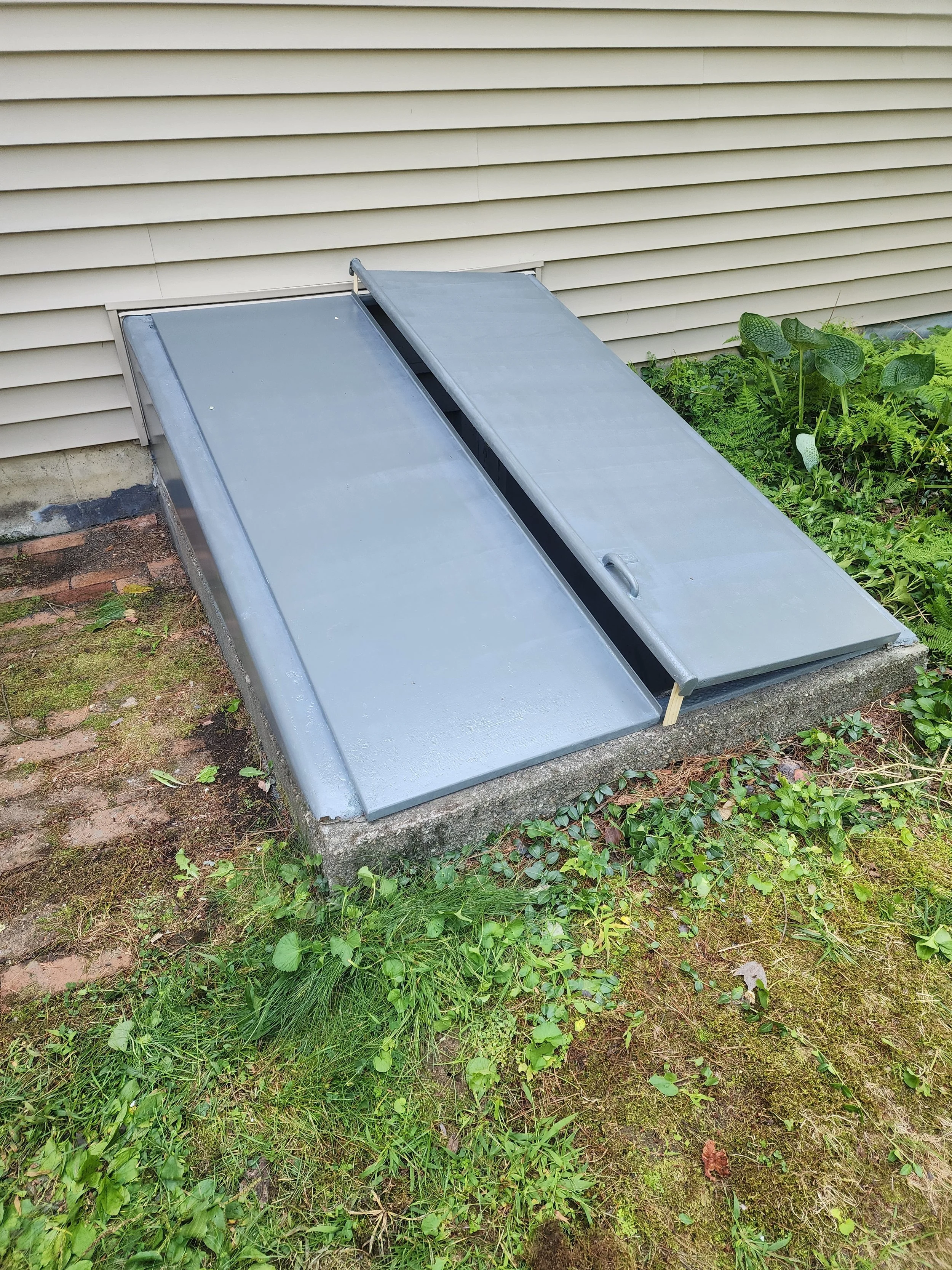 A metal emergency exit ramp with a sliding cover, installed outside next to a beige house with horizontal siding and surrounded by green grass and plants.