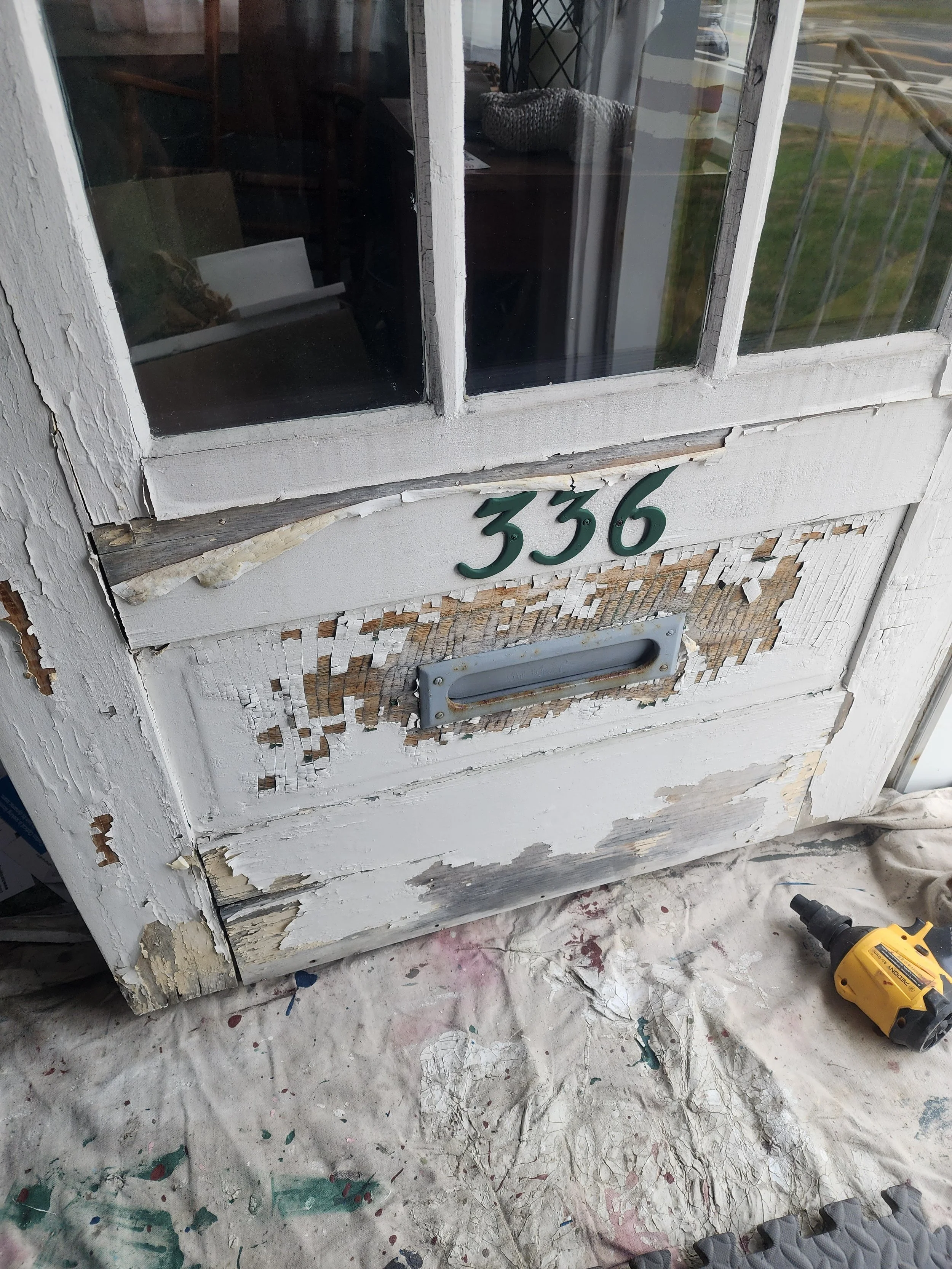 The front door with peeling white paint and a metal mail slot, displaying house number 336, with visible damage and chipping wood, and a power drill on the floor nearby.