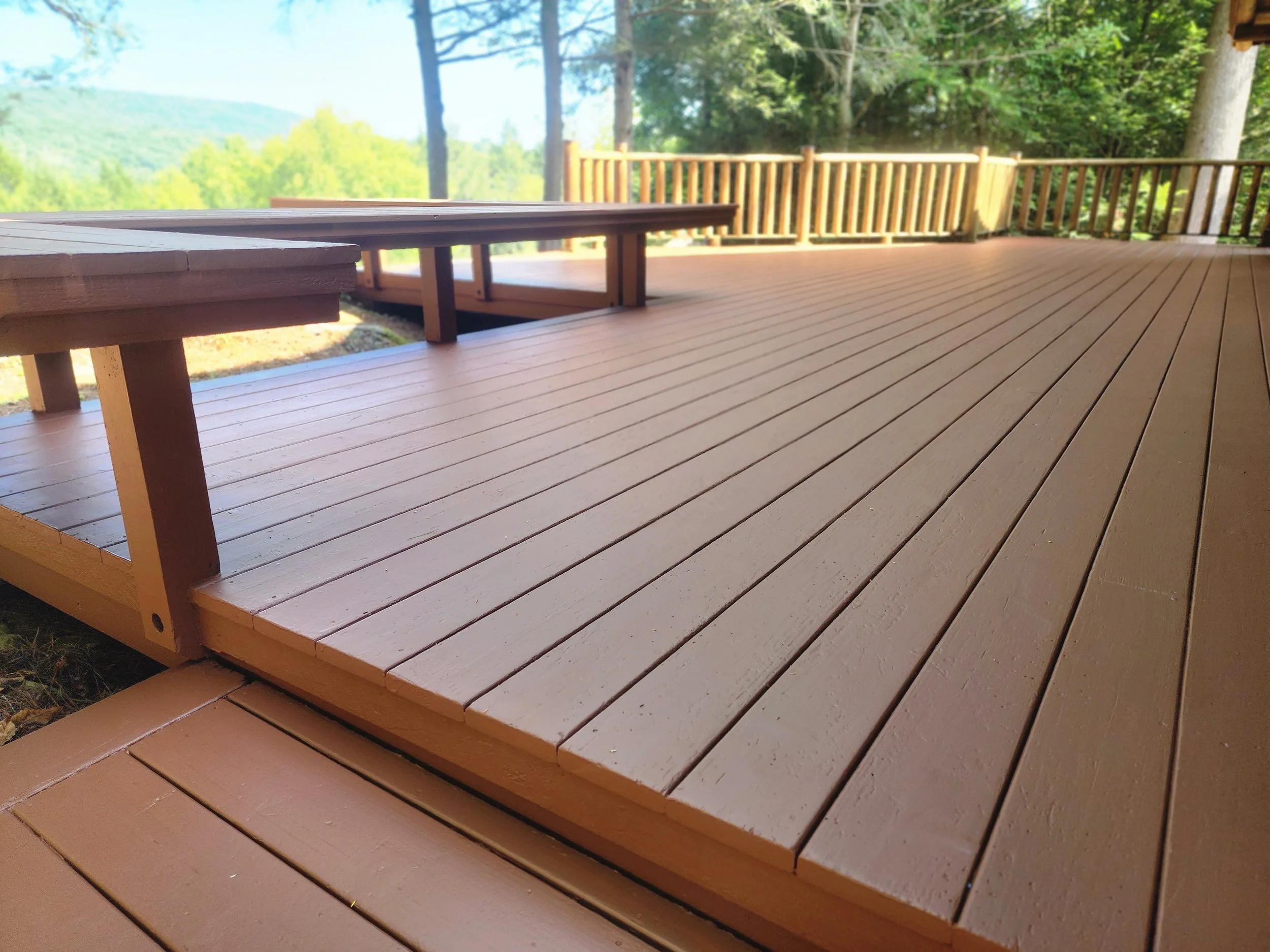 View of a wooden deck with a bench and railing, surrounded by trees and mountains in the background, during daytime.
