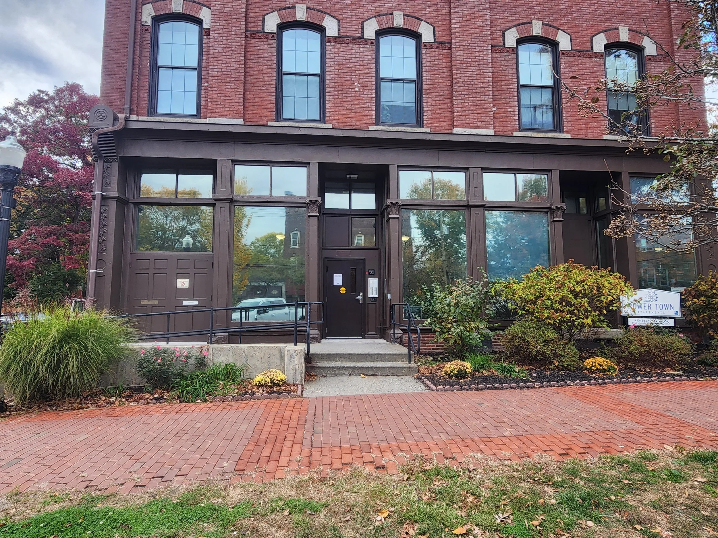 Brick building with large glass windows and a door, surrounded by a small garden with shrubs and flowers, on a brick-paved sidewalk.