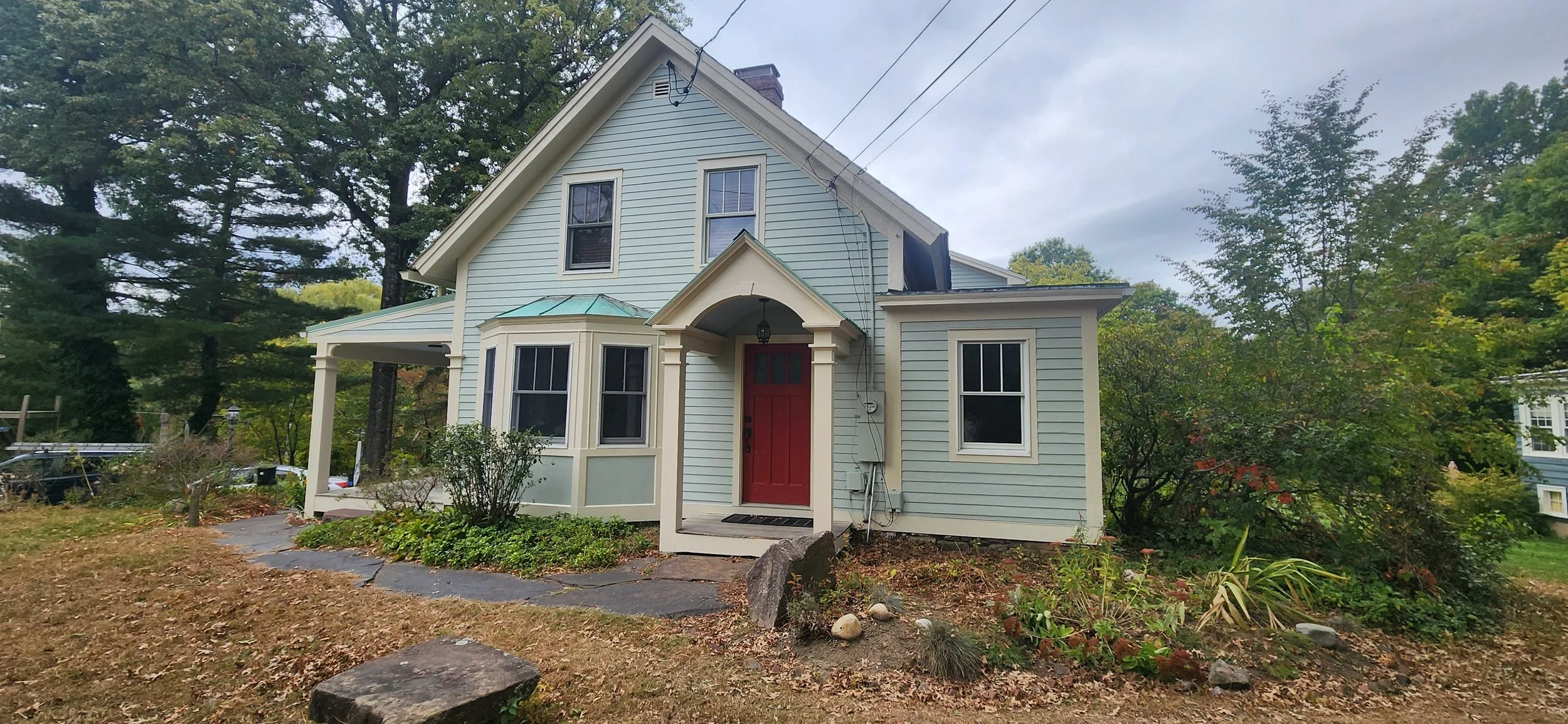 A two-story light blue house with white trim, a red front door, and a small porch area. There is a bay window on the front with a small roof, and several trees and shrubs surrounding the house. The yard has patches of brown grass and rocks, with a wa