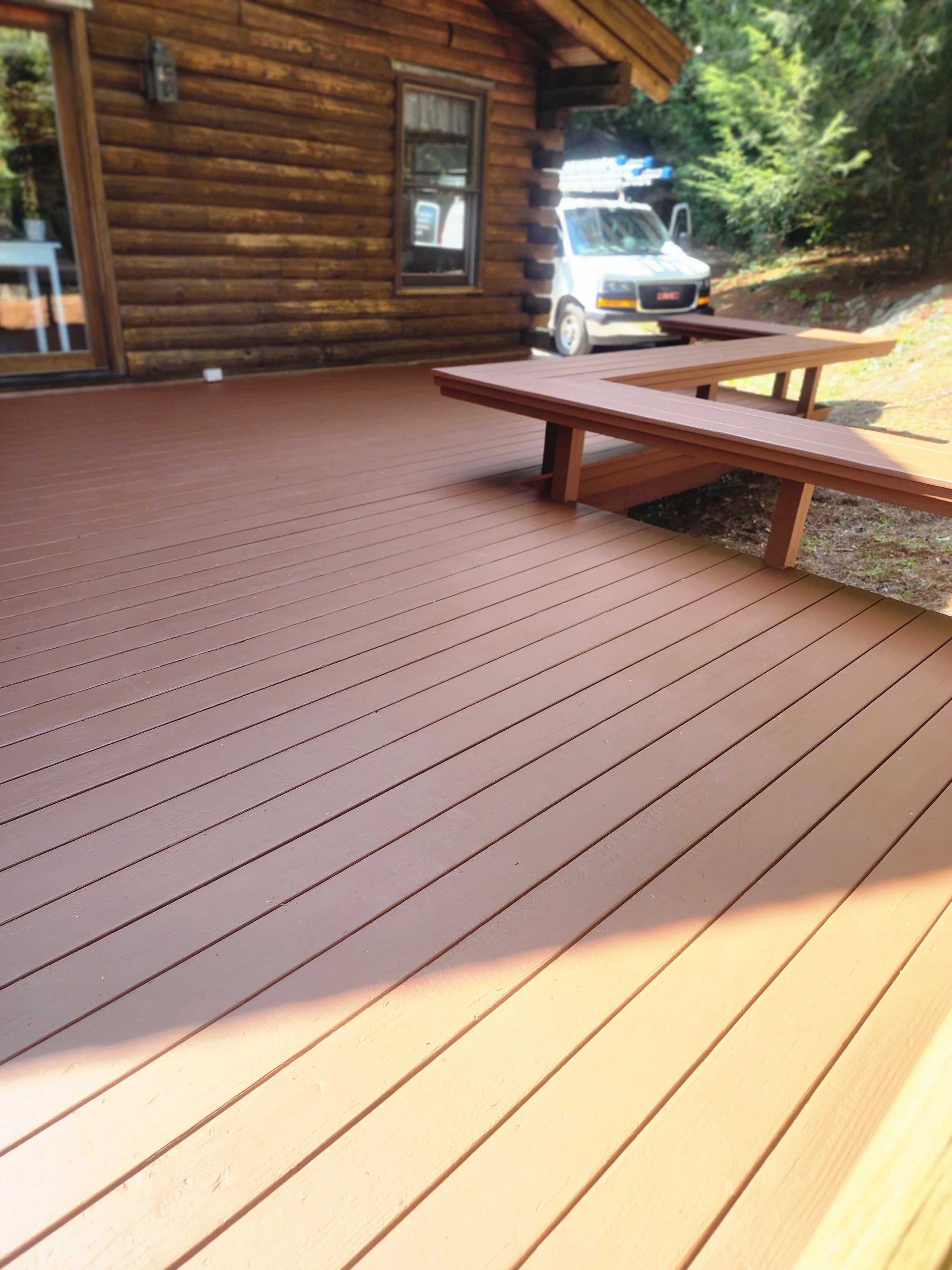 Wooden deck attached to a log cabin, with built-in benches, and a van parked in the yard.