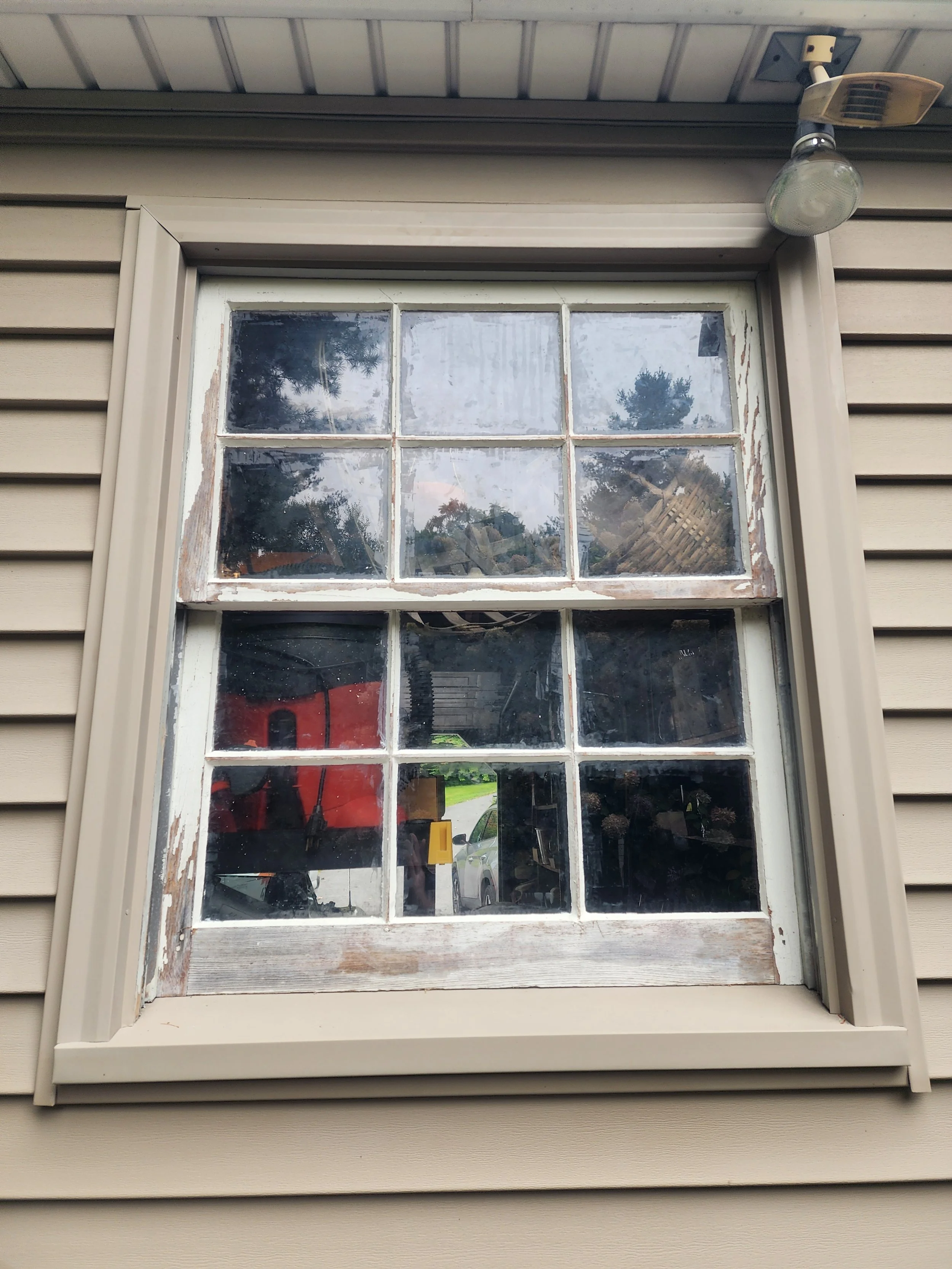 A weathered double-hung window with six panes in a beige house, showing signs of peeling paint and age. The window is set in beige siding beneath a metal overhang, with trees, a red object, and a car visible outside.