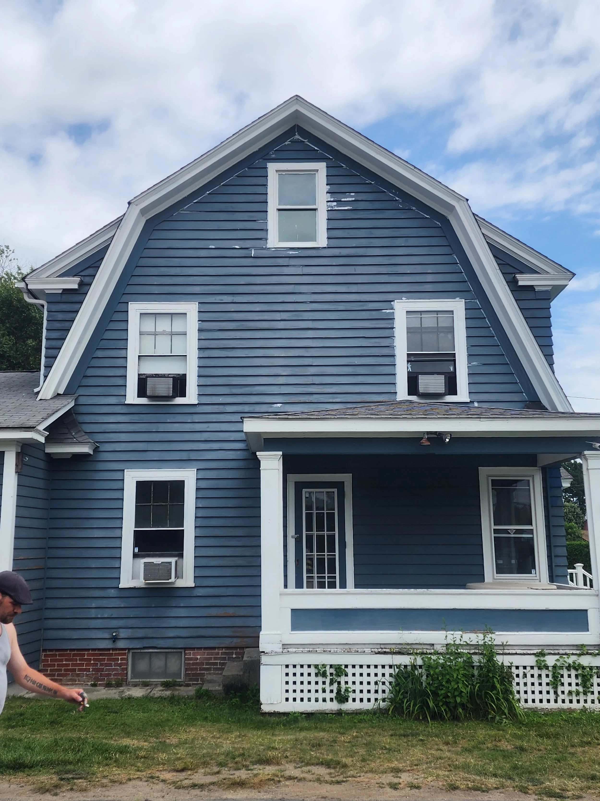 Blue three-story house with white trim, front porch, and windows with air conditioning units. Part of a person is visible on the left side.