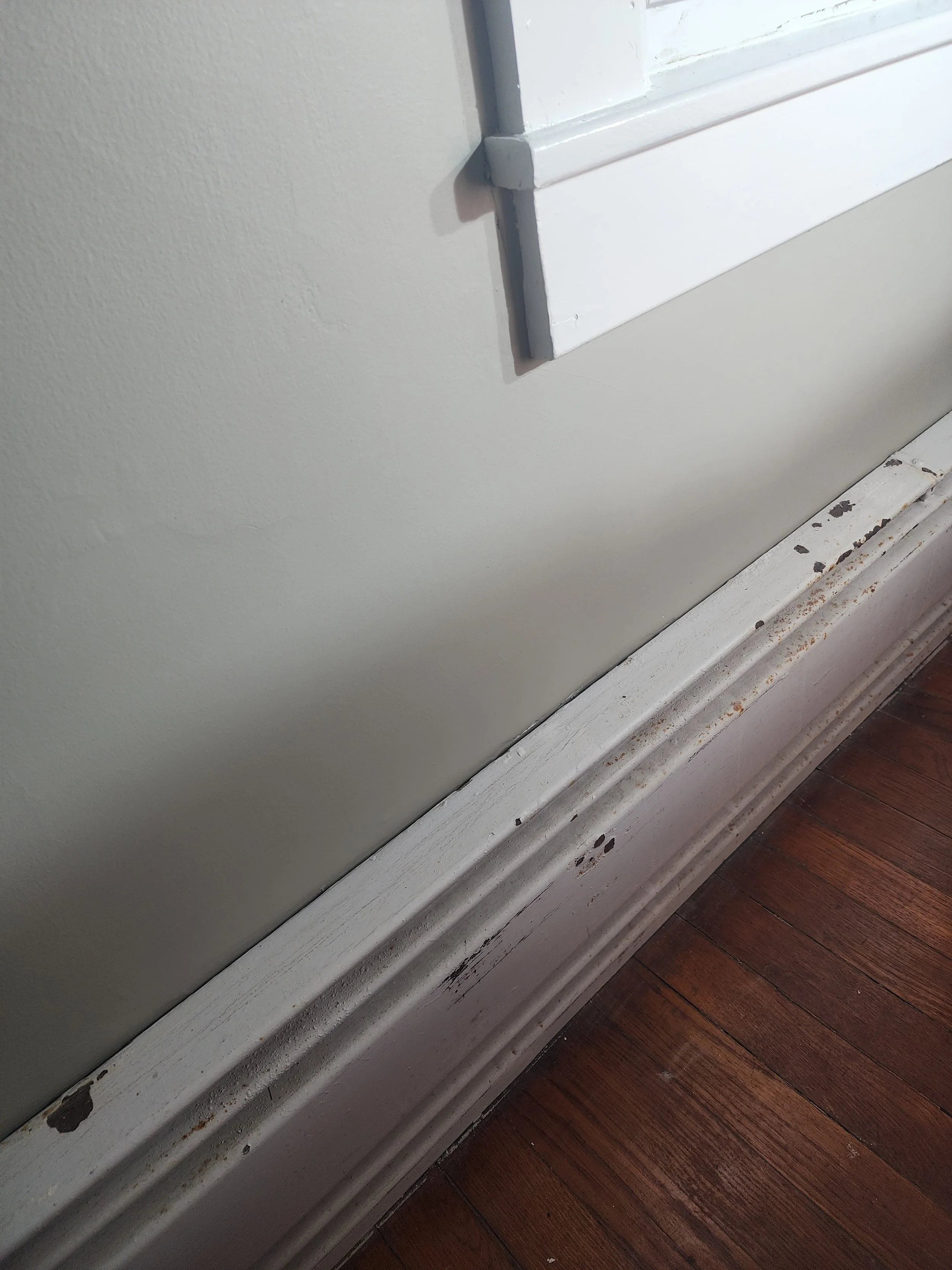 Close-up photo of a baseboard heater with chipped white paint and rust, situated on a dark hardwood floor.