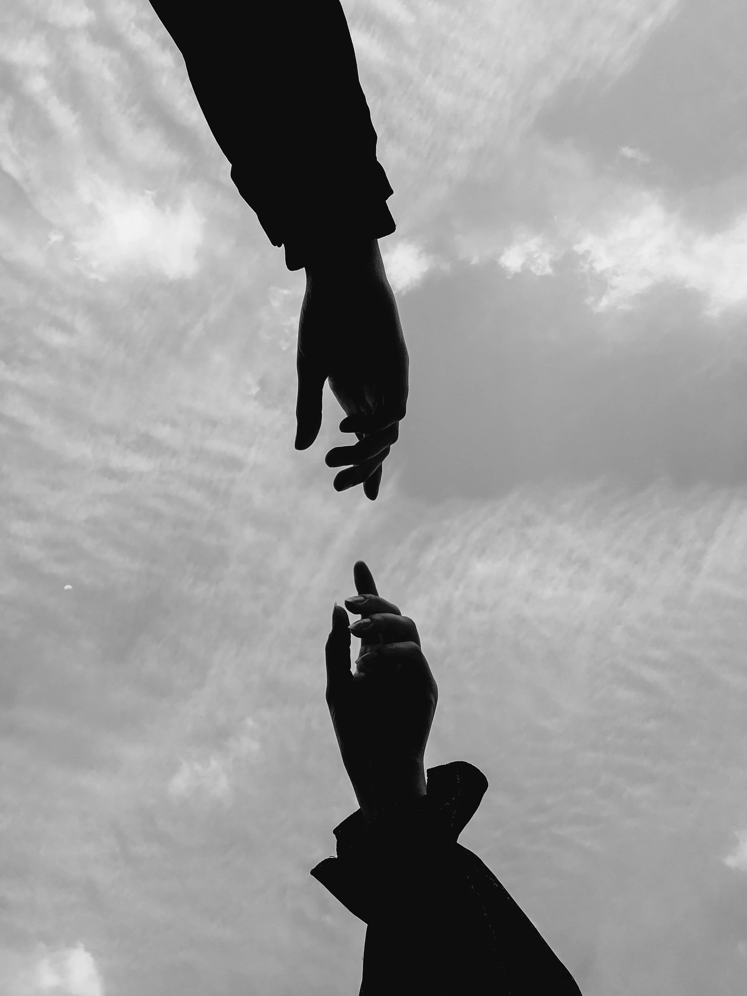 Silhouettes of two hands reaching towards each other against a cloudy sky.
