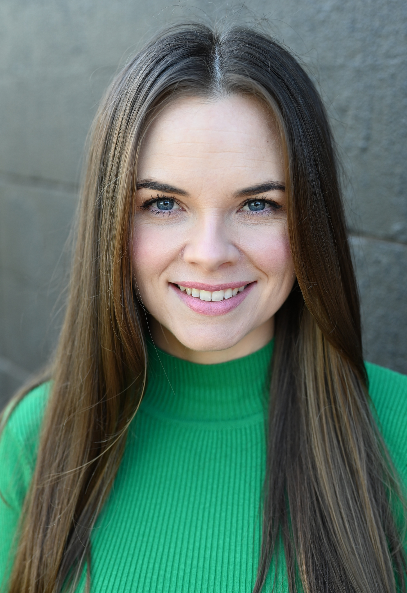 Close-up of a smiling woman with long brown hair, blue eyes, wearing a green top, standing against a gray wall.
