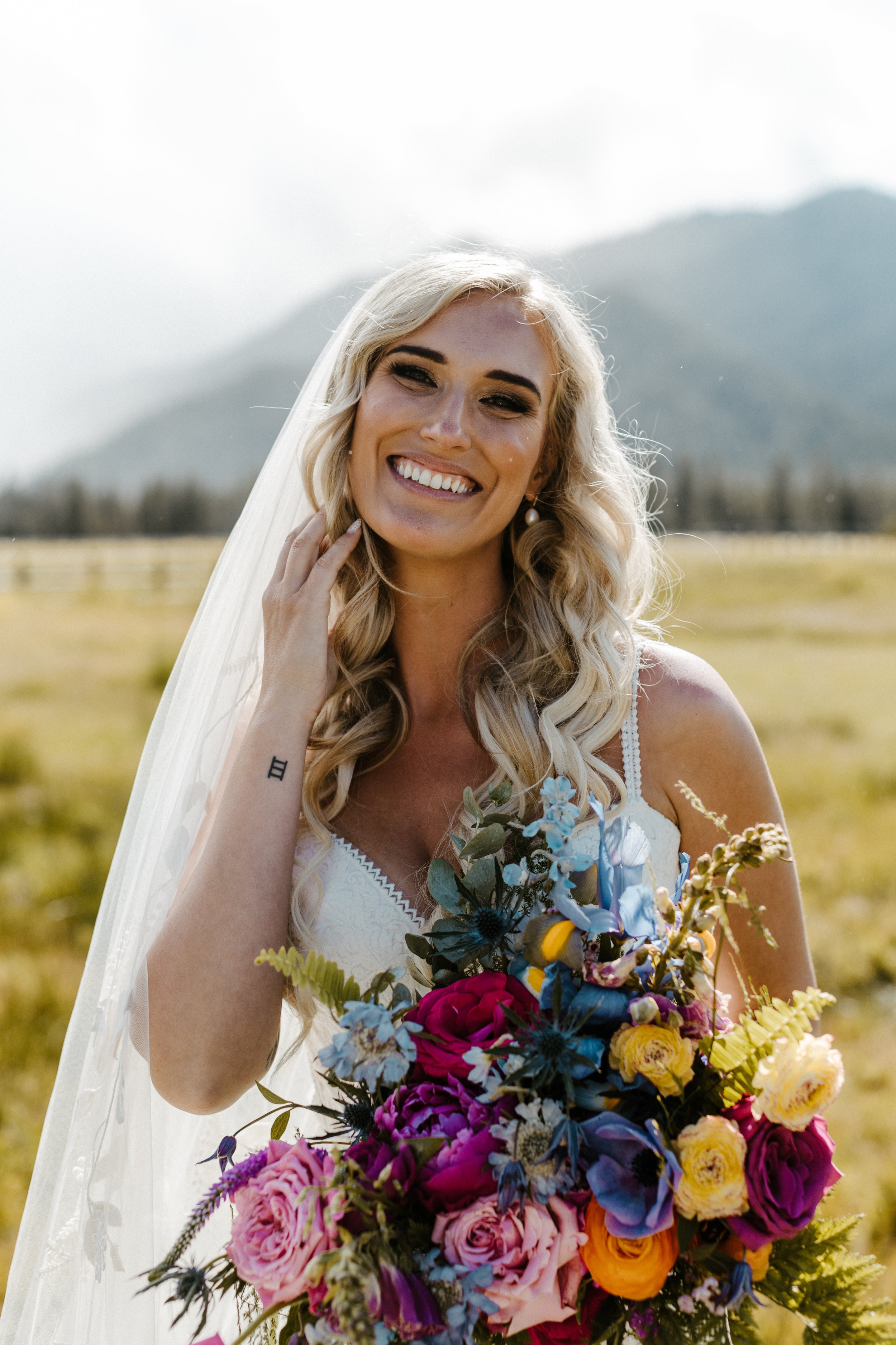 Bride smiling in wedding dress with colorful bouquet, standing outdoors with mountains and fields in the background.