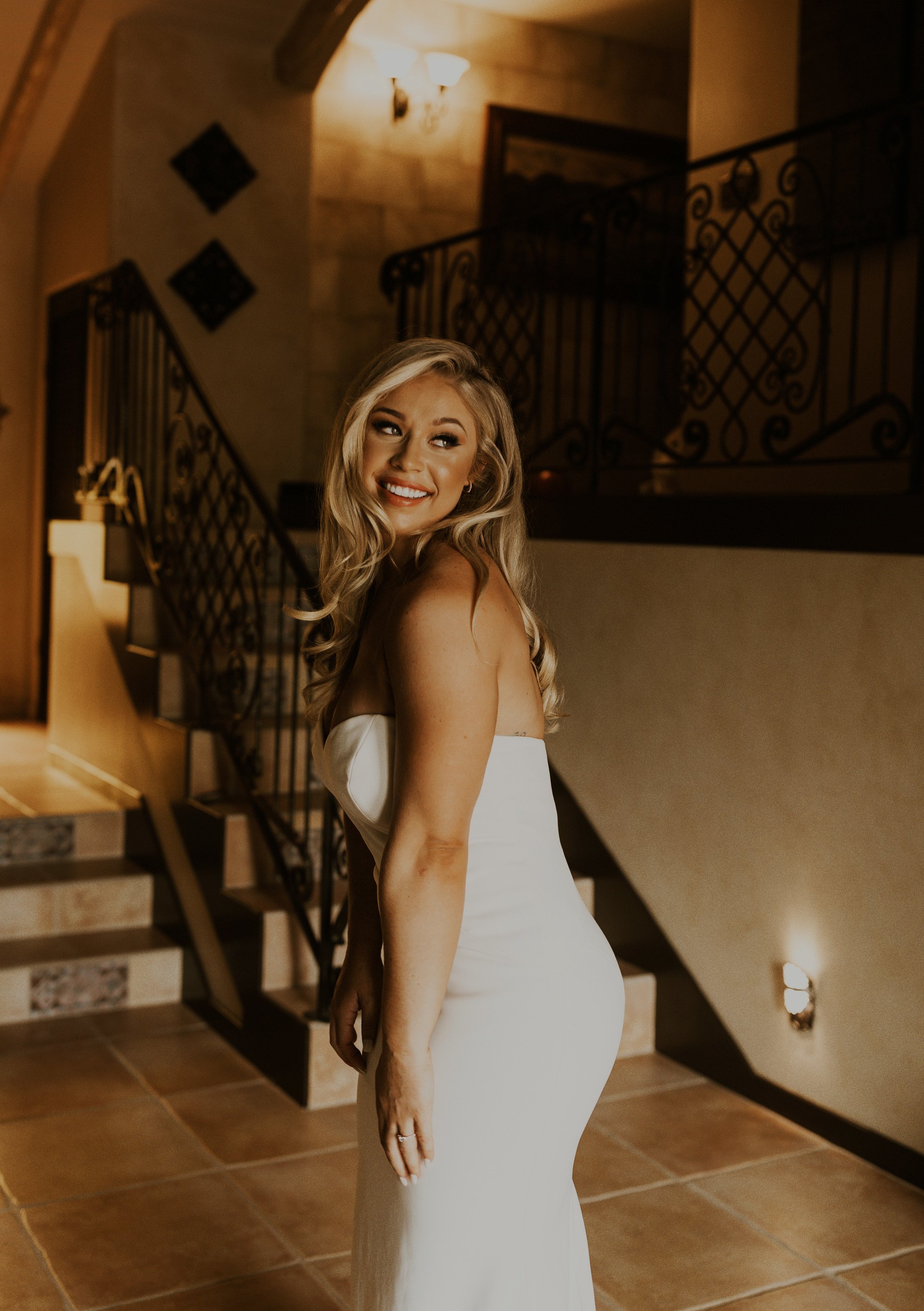 Woman in a white dress smiling indoors near staircase.
