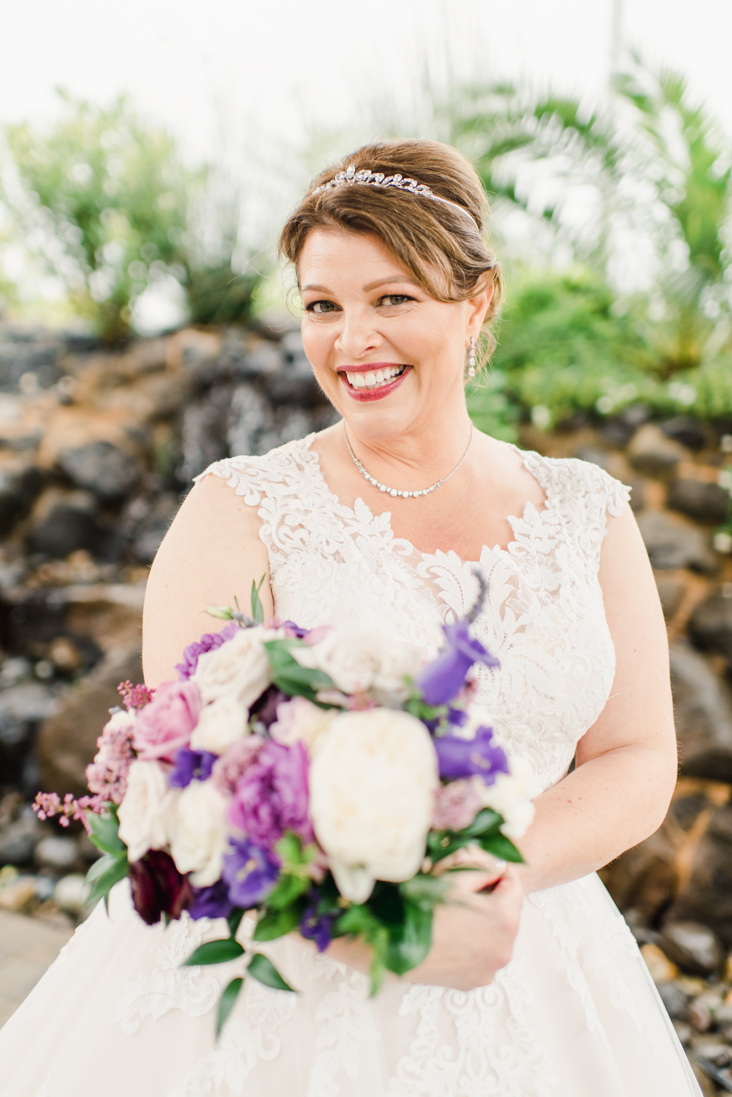 Smiling bride in a white lace wedding dress holding a colorful bouquet of flowers, outdoors with greenery in the background.