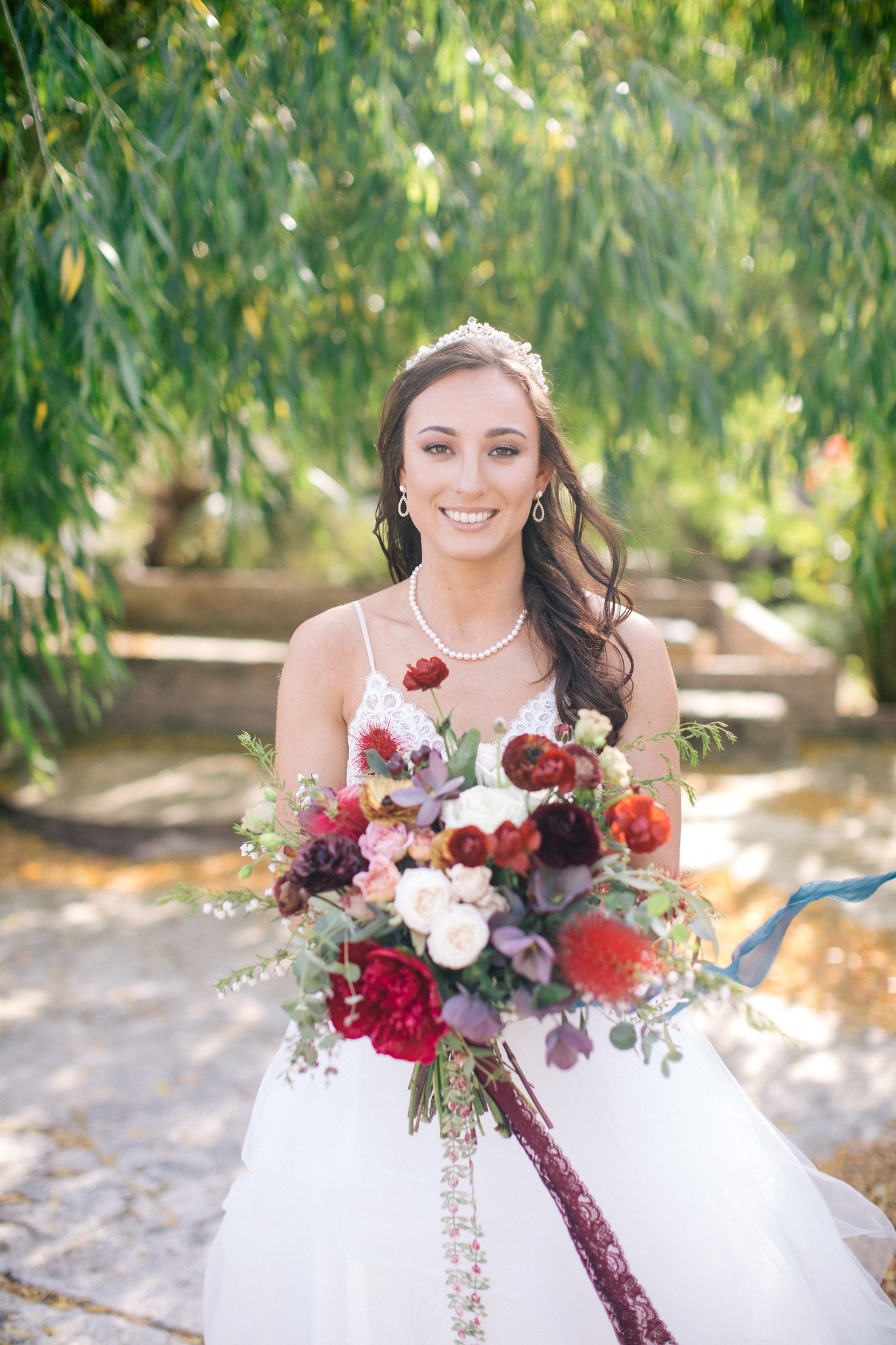 Bride in white dress holding a colorful bouquet with red and pink flowers, standing outdoors with green foliage in the background.