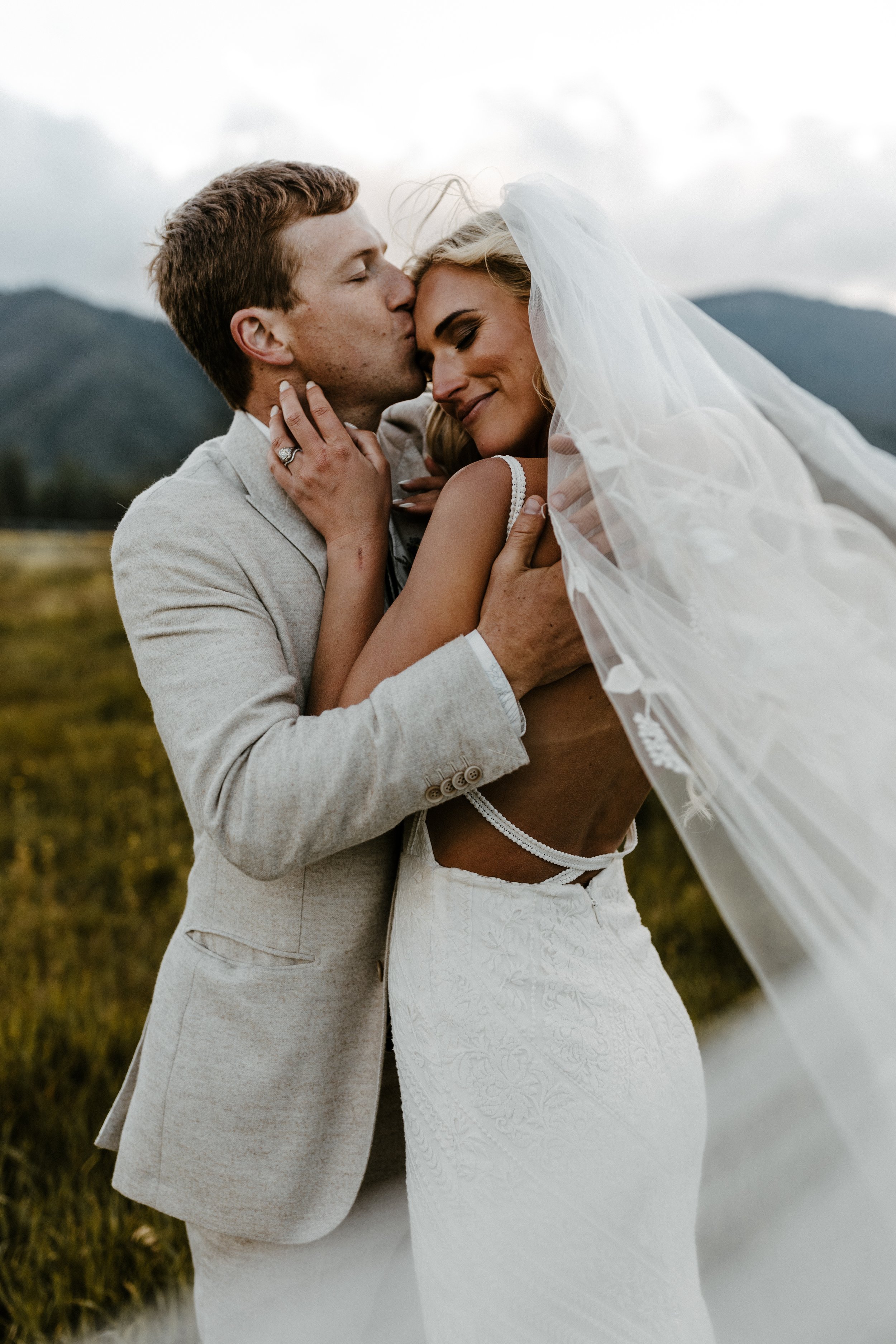 A couple in wedding attire embracing each other outdoors, with mountains in the background. The woman is wearing a white wedding dress and veil, while the man is in a light-colored suit.