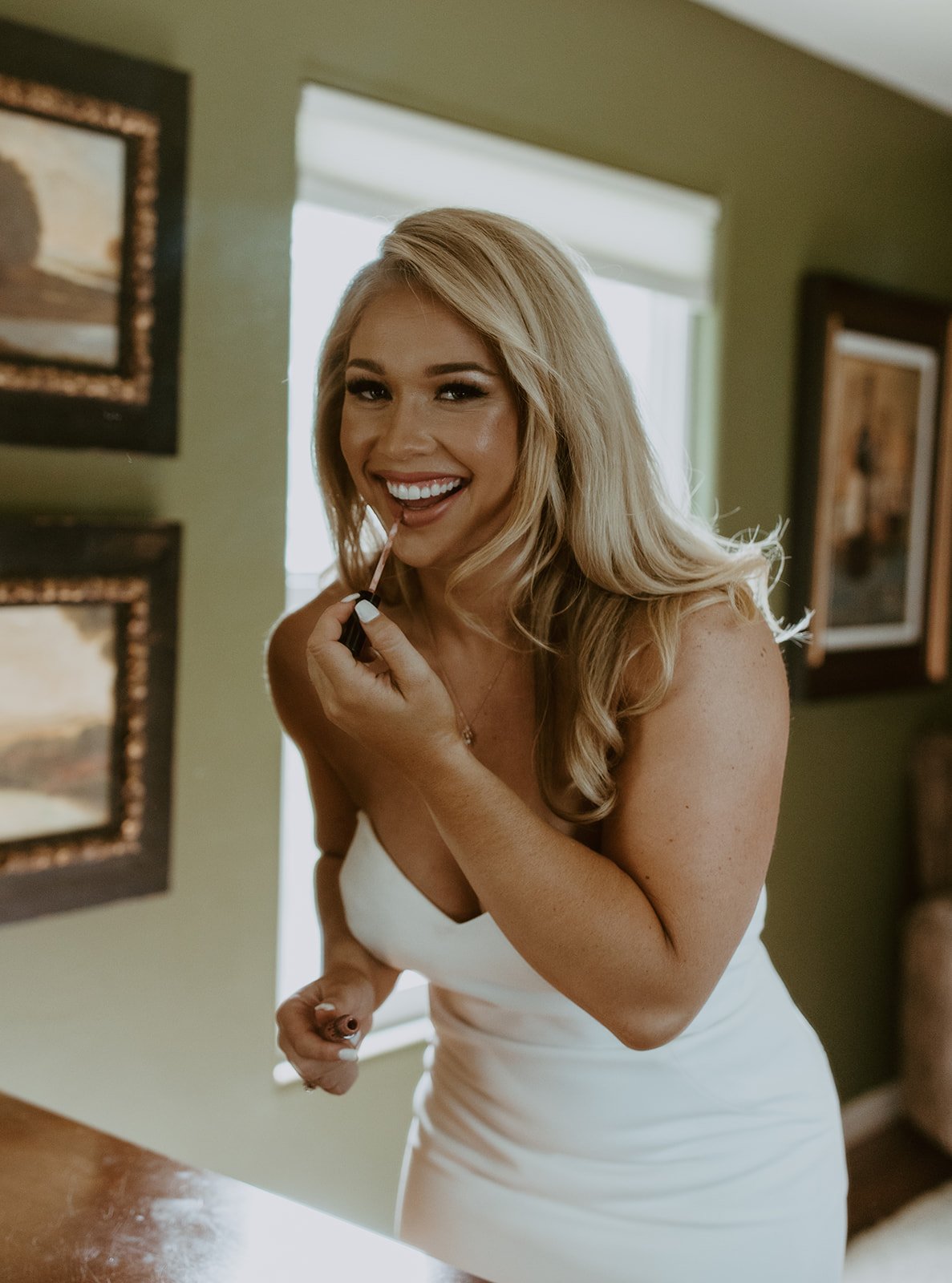 Woman in white dress applying lip gloss, smiling.