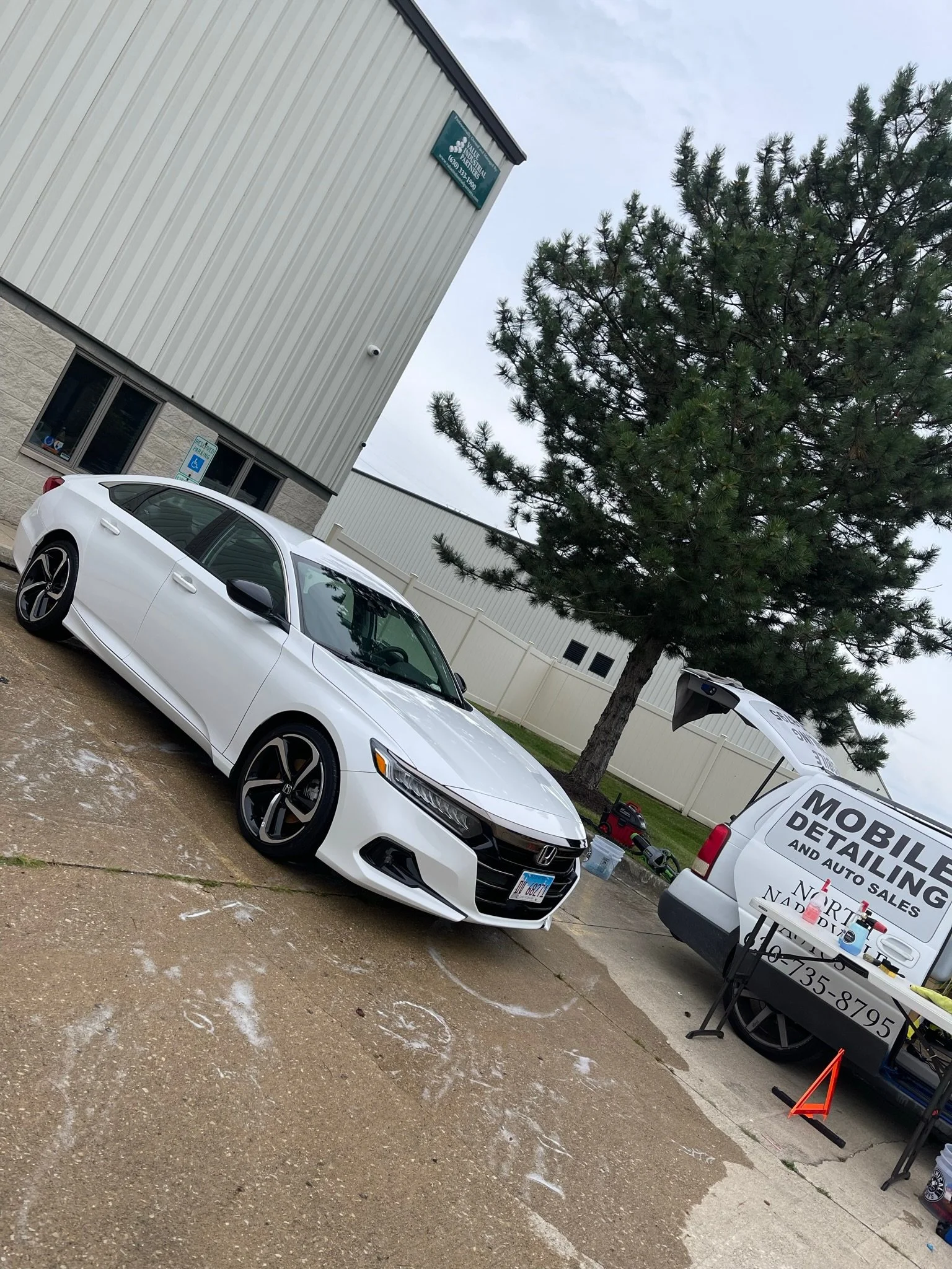 A white Honda sedan parked outdoors near a tree, with a mobile detailing van and a table with cleaning supplies nearby.