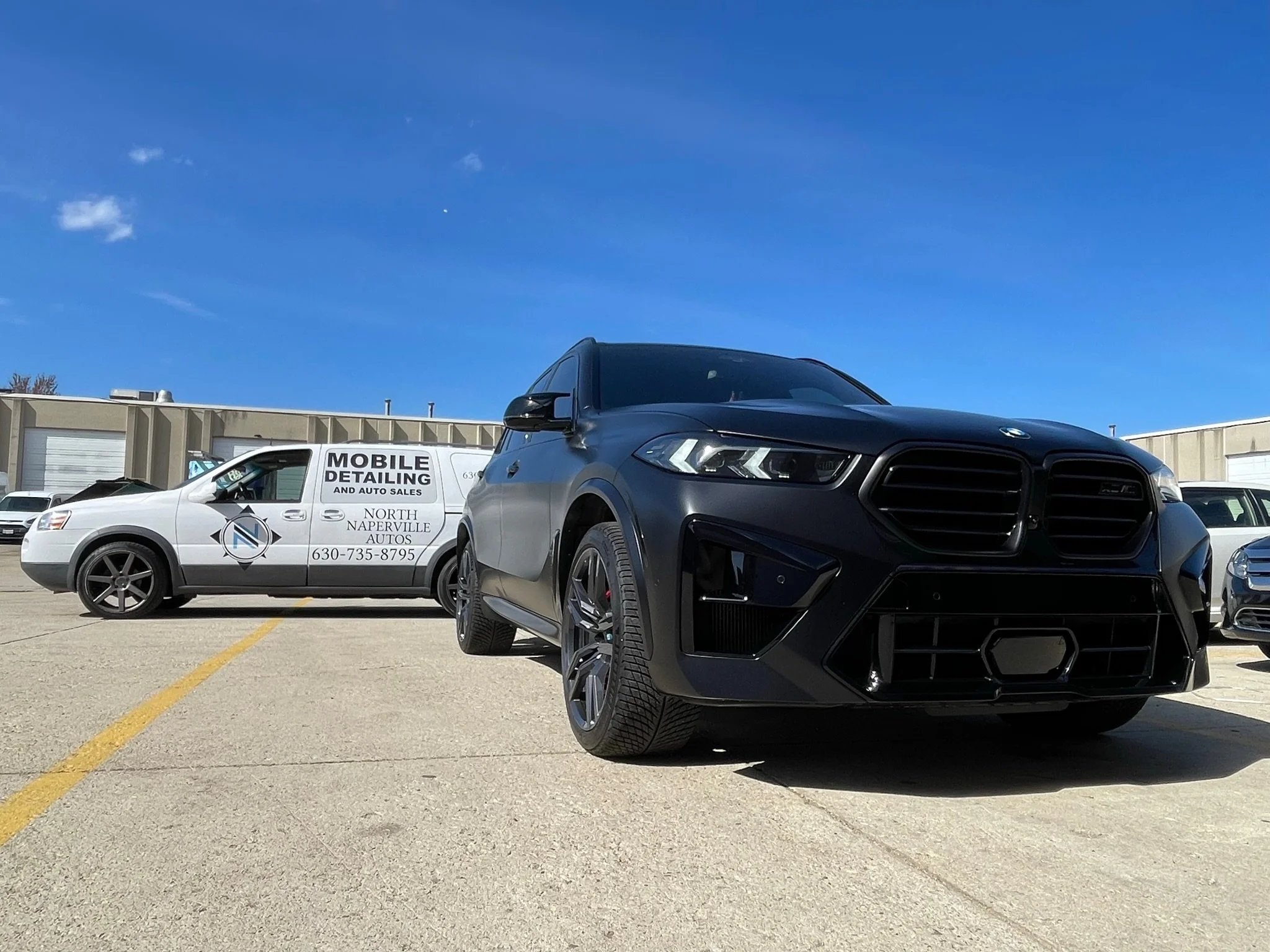 Black luxury SUV and white vehicle with car detailing business signage parked in an outdoor lot under a clear blue sky.