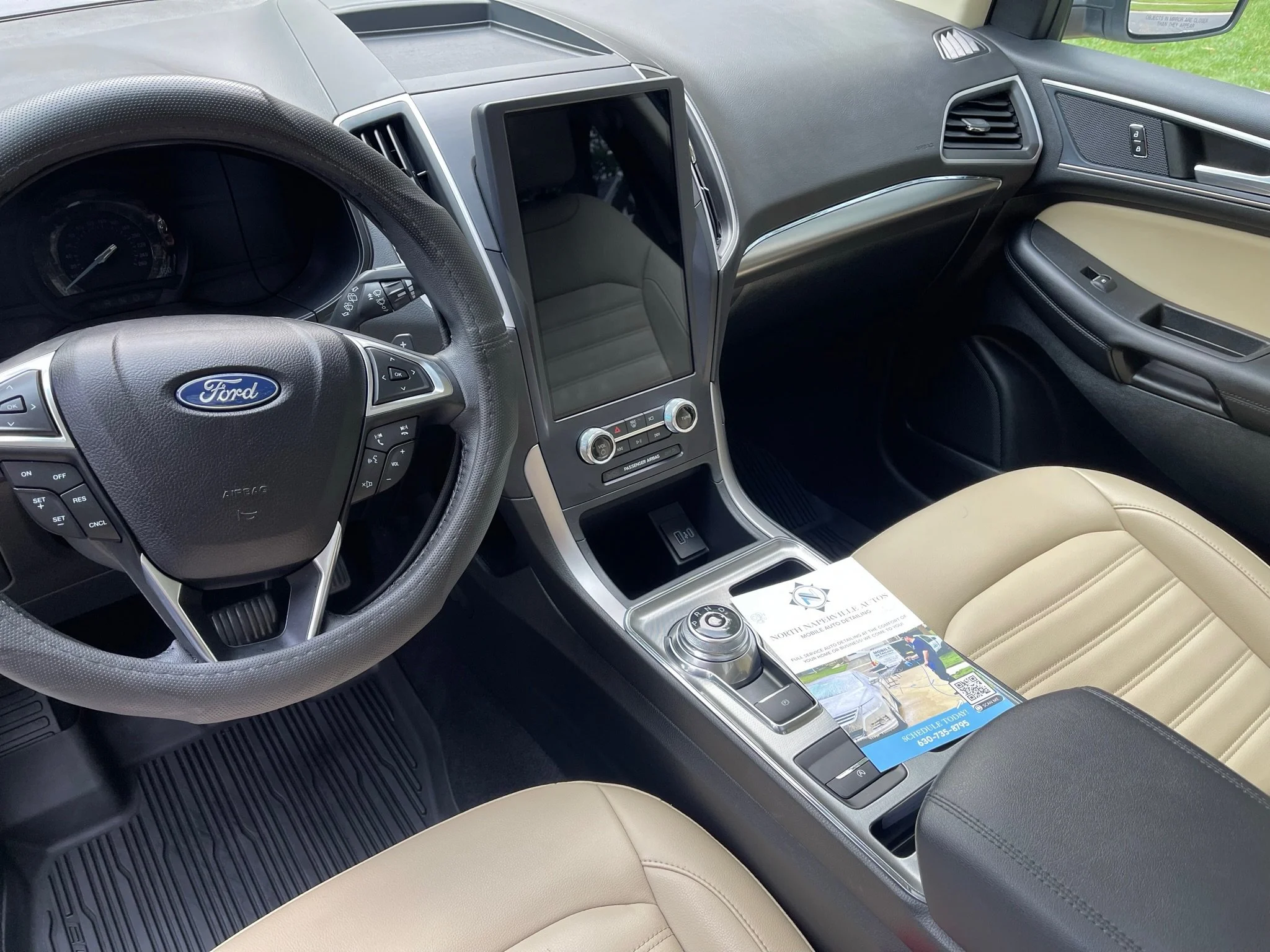 Interior of a Ford vehicle showing steering wheel, center console with large touchscreen, gear selector, and beige leather seats.