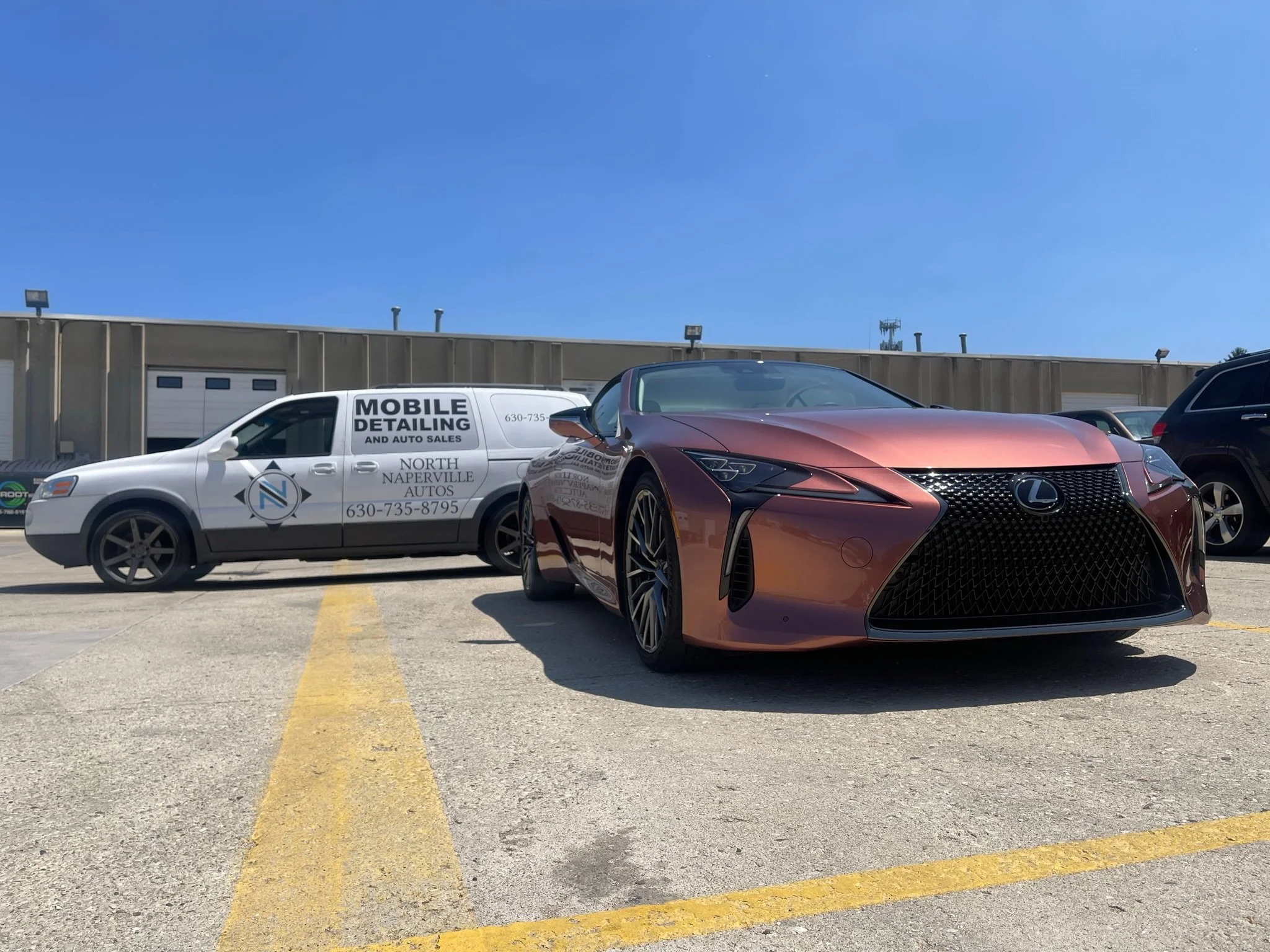 A red Lexus sports car parked in a parking lot next to a white mobile detailing van with black text and contact information.