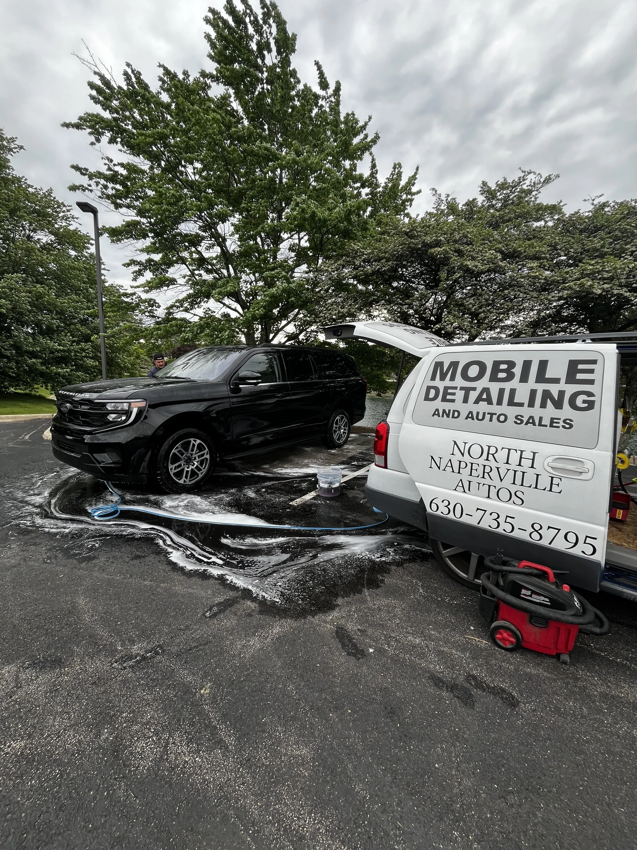 A black SUV being washed with soap and water outdoors next to a white mobile detailing van with advertising text, including a phone number and location in North Naperville, Illinois. The van's side door is open.
