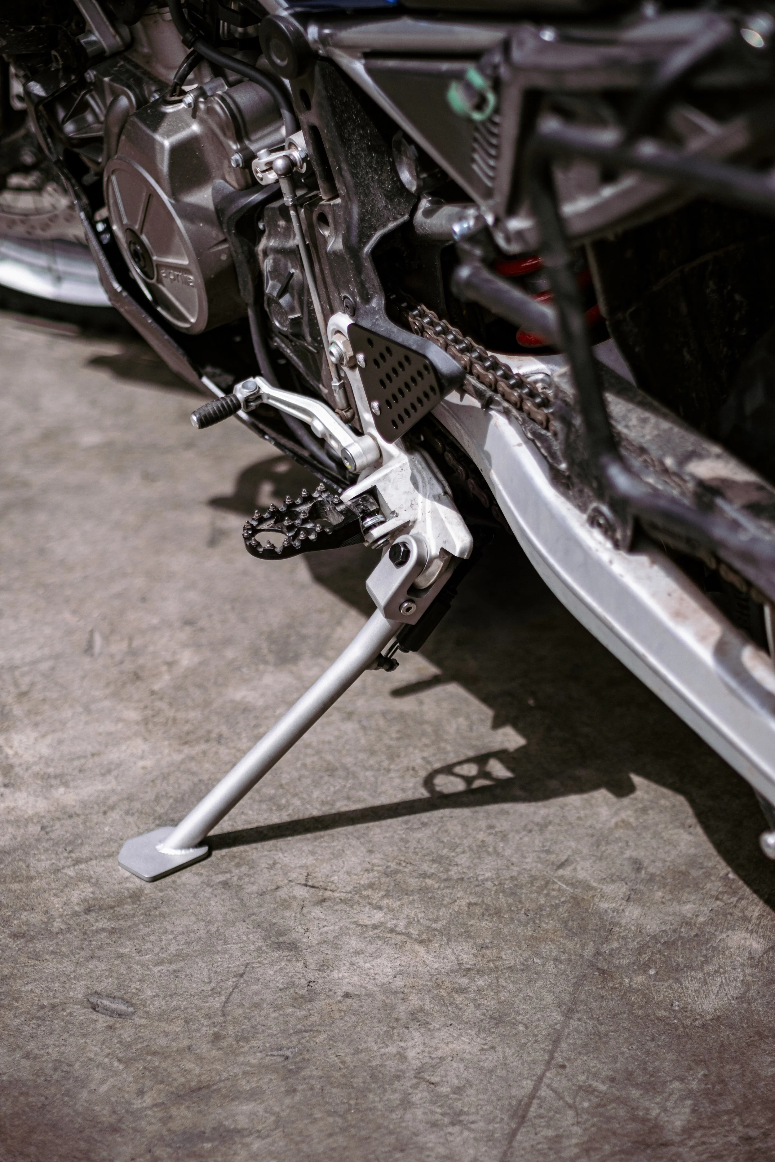 Close-up of a motorcycle's engine and foot pegs, showing the side stand on a concrete surface.