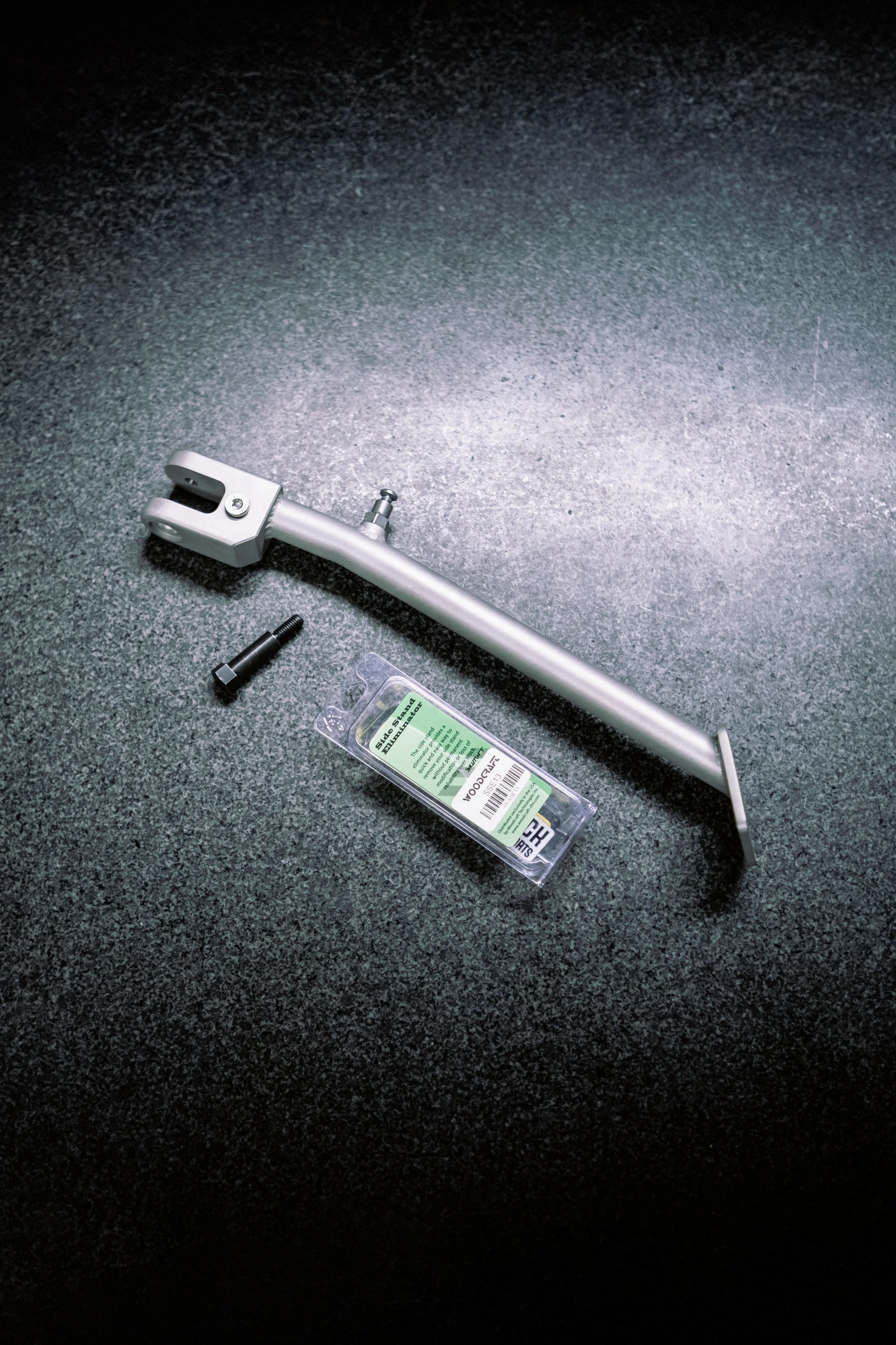 A set of motorcycle or bicycle repair tools on a dark textured surface, including an axle or fork tube, a bolt, and a small plastic package of parts.