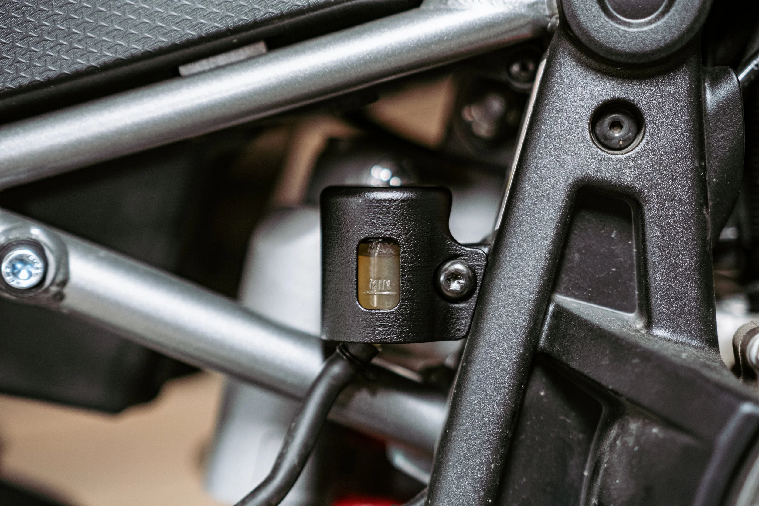 Close-up of a mechanical part on a motorcycle, showing a black component with a small fluid level window and surrounding metal framework.