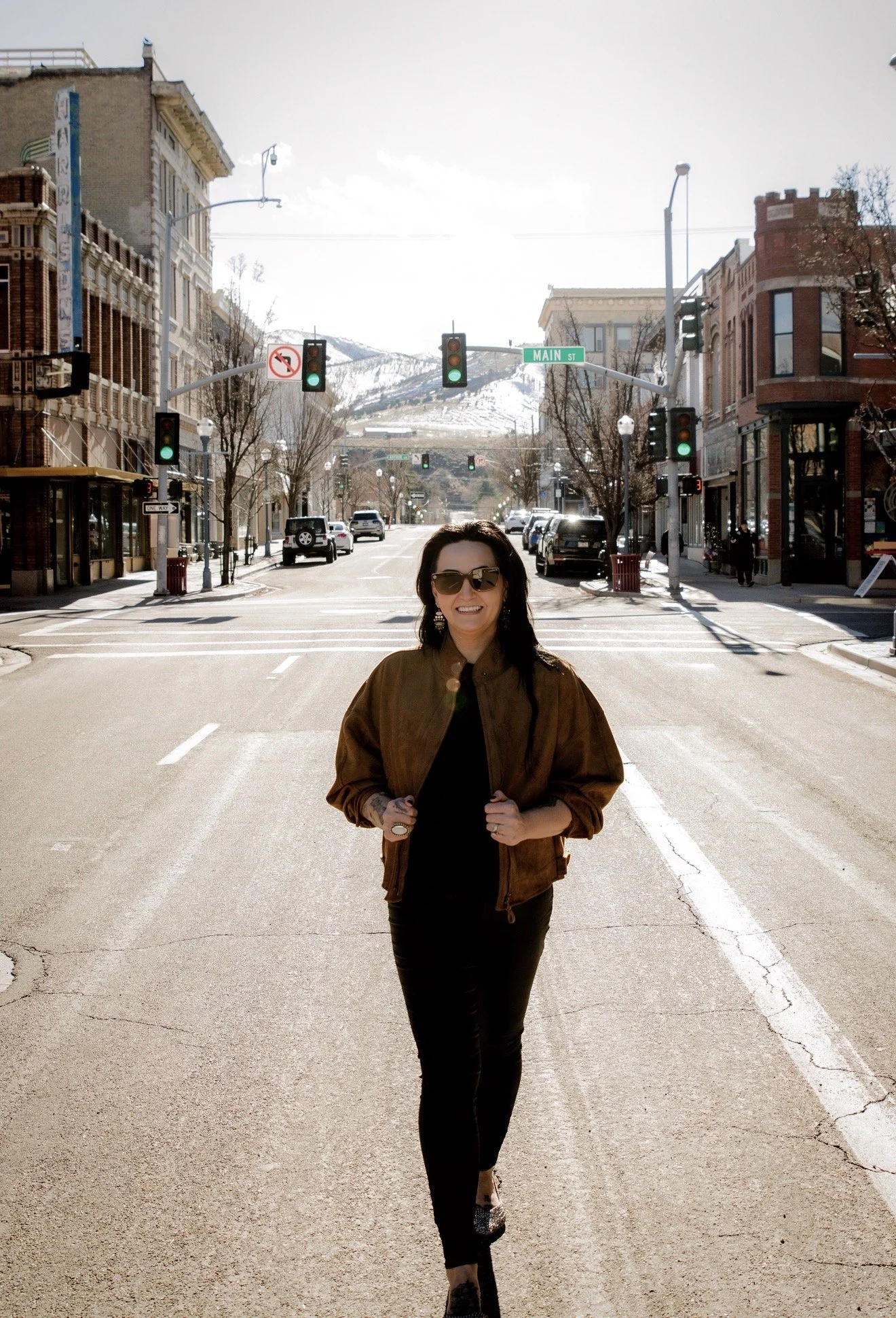 A woman standing in the middle of a city street with buildings on both sides, wearing sunglasses, a brown jacket, and black pants, smiling at the camera in bright daylight. There are cars parked along the street and snow-capped mountains in the background.