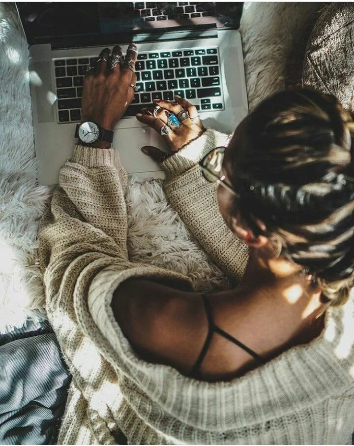 A person lying on a bed with a fluffy blanket, typing on a laptop. They are wearing a knitted sweater, multiple rings, bracelets, glasses, and have a braided hairstyle.