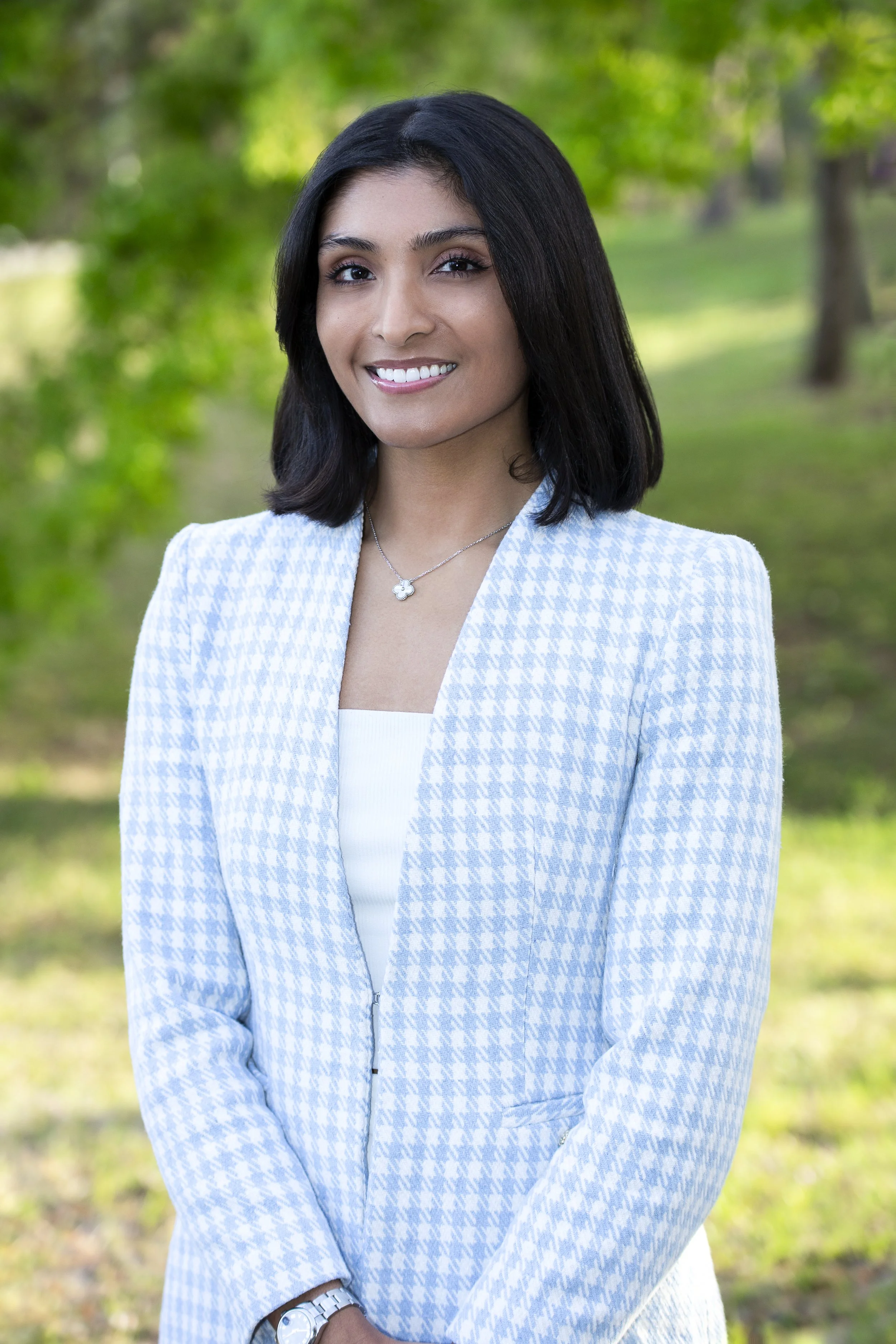 A woman standing outdoors in front of a blurred green background, wearing a light blue and white houndstooth blazer over a white top. She has shoulder-length dark hair, is smiling, and is wearing a delicate necklace and wristwatch.
