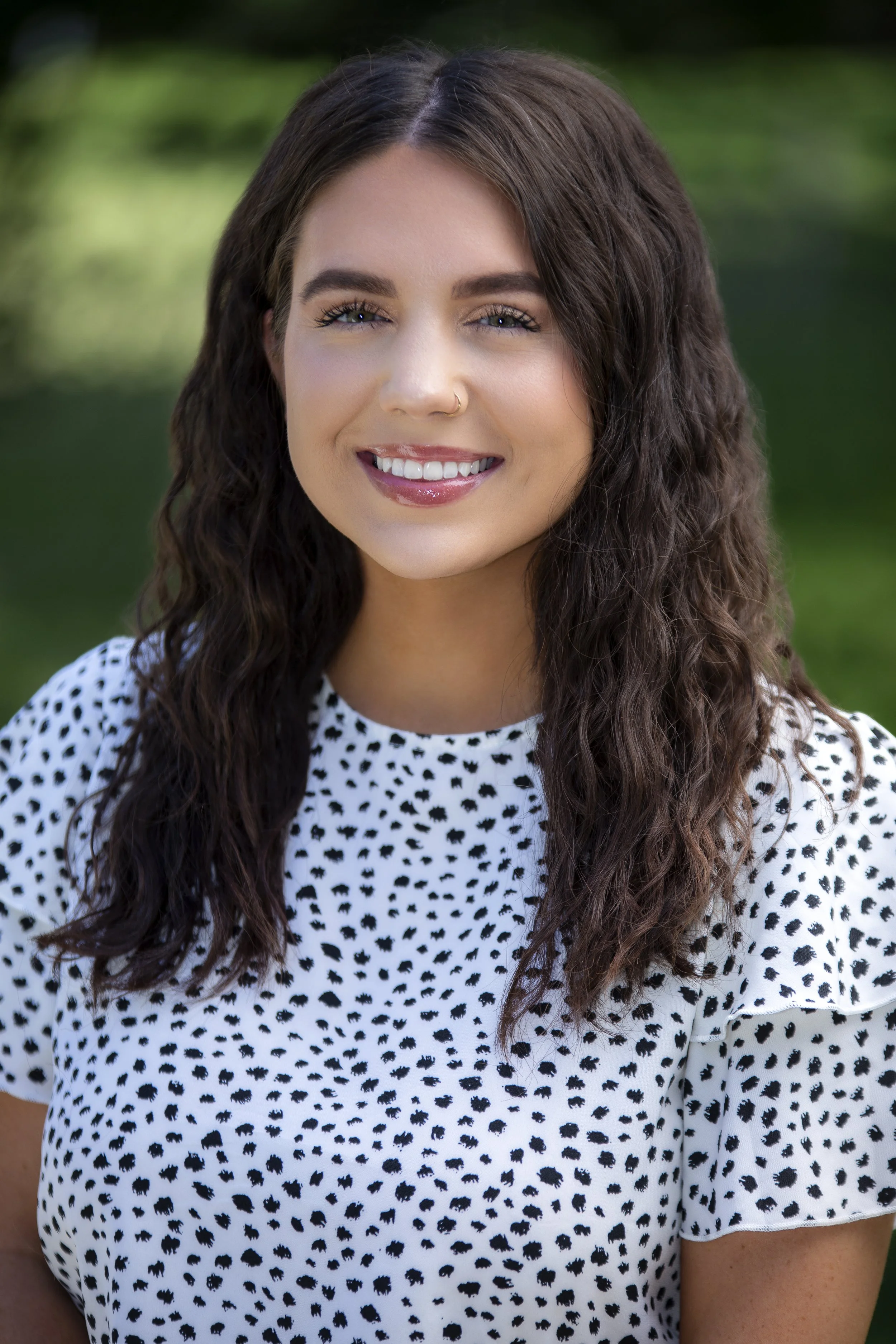 A woman standing outdoors with a blurred green background, smiling at the camera. She has long, wavy dark brown hair, is wearing a white blouse with a black abstract dot pattern, and has a small nose ring on her left side.
