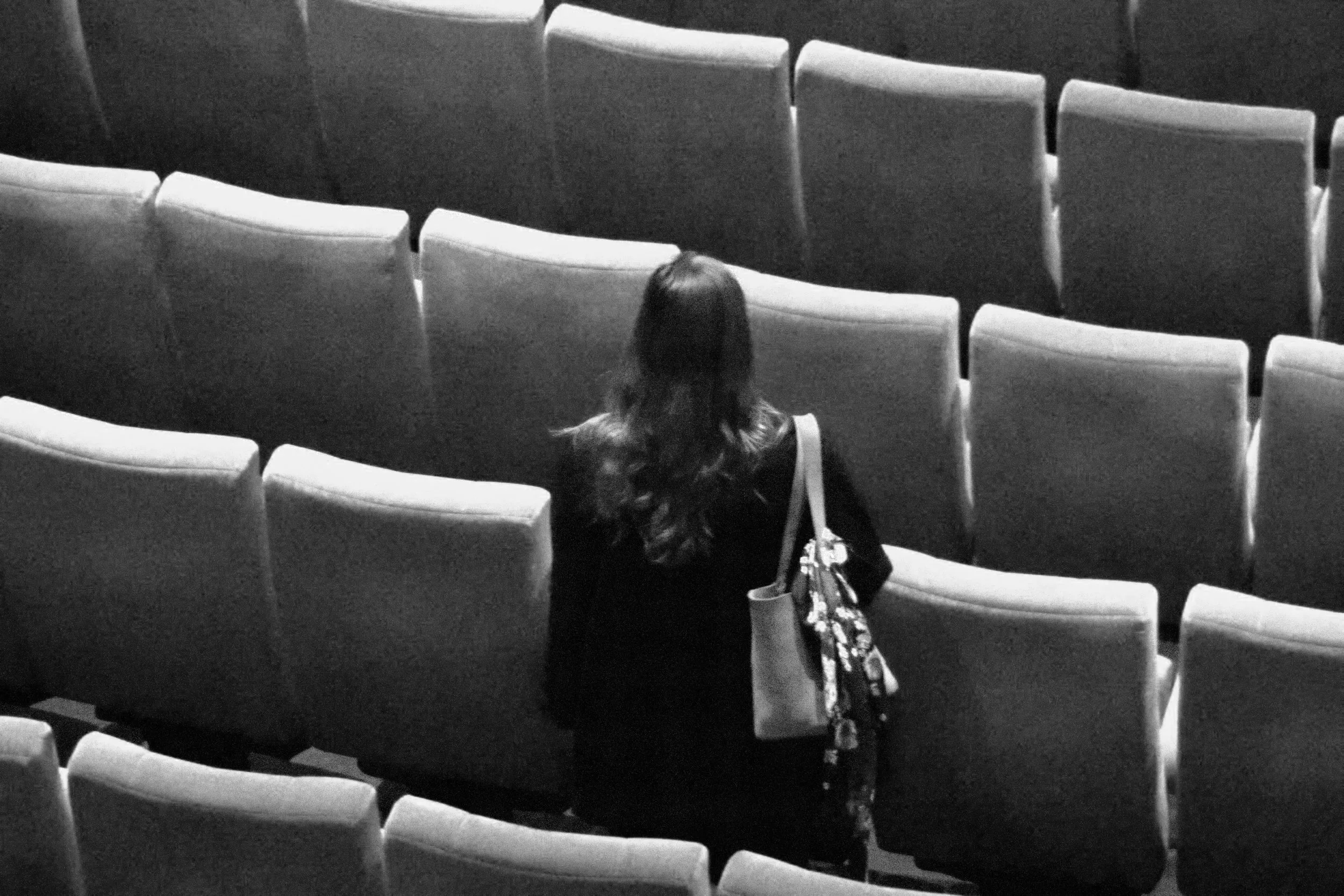 A woman with long hair, carrying a tote bag and a purse, is seated alone in a mostly empty theater or auditorium with rows of empty seats, viewed from behind.