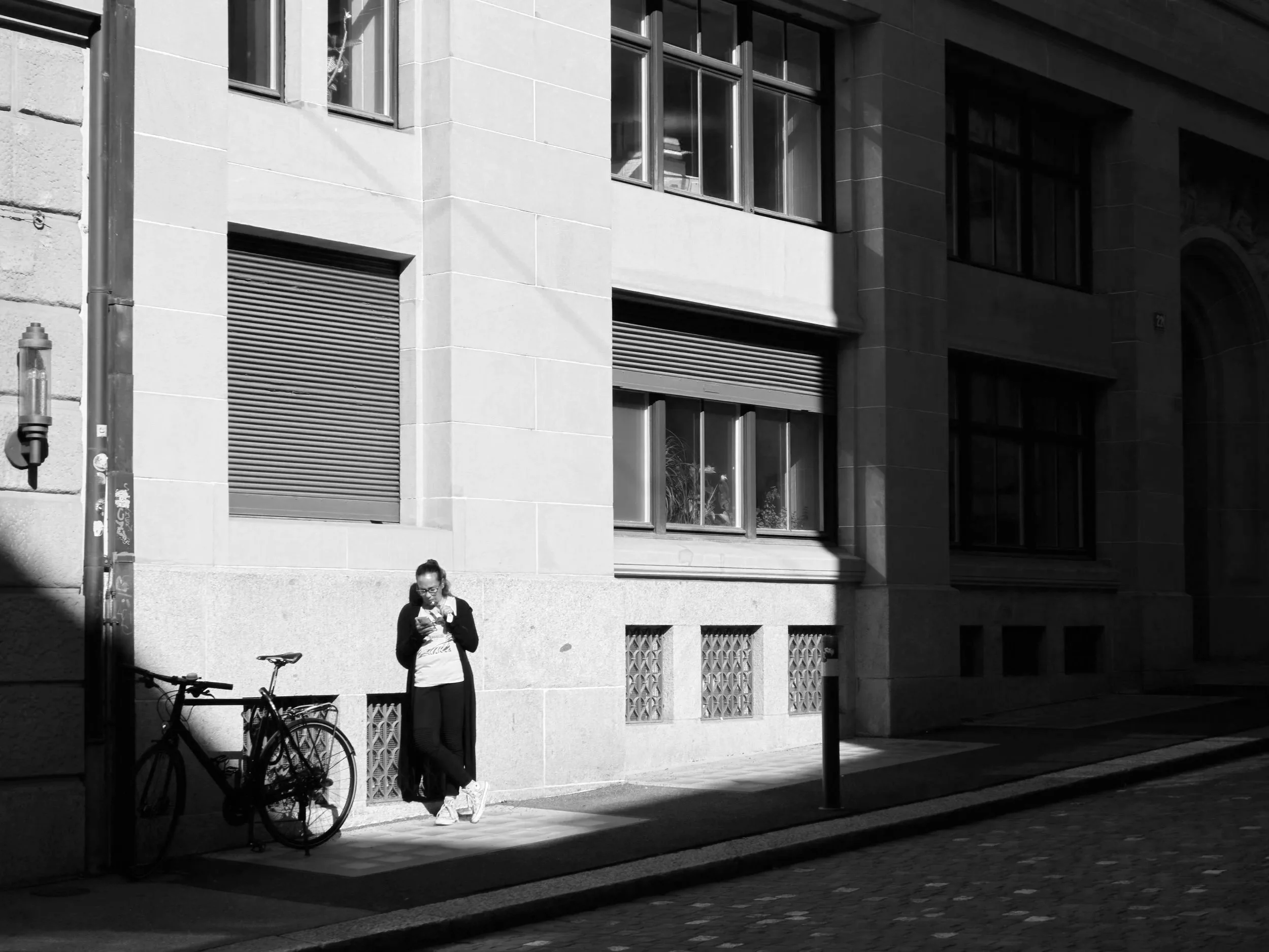 A woman standing on a city sidewalk with a bicycle, looking at her phone, with a building's exterior wall and windows in the background, partially shaded by a shadow.