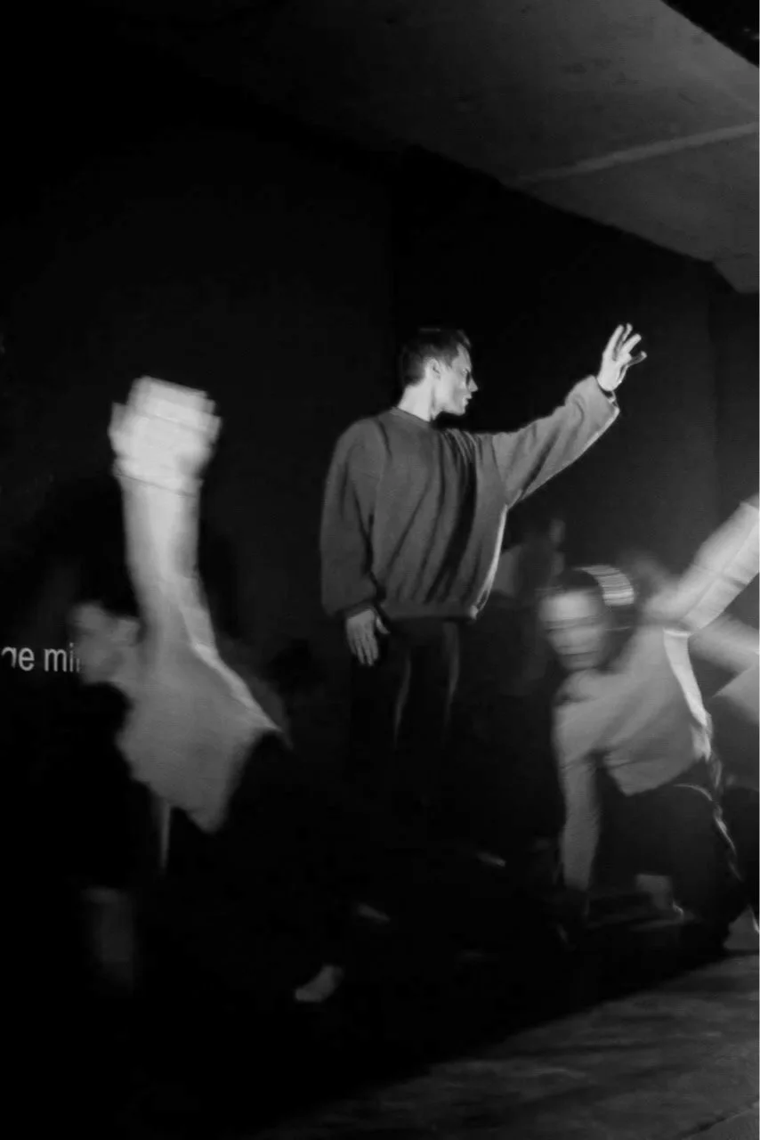 A black-and-white photo of a person on stage reaching out with their right hand, with other people in motion around them, possibly during a performance or speech.