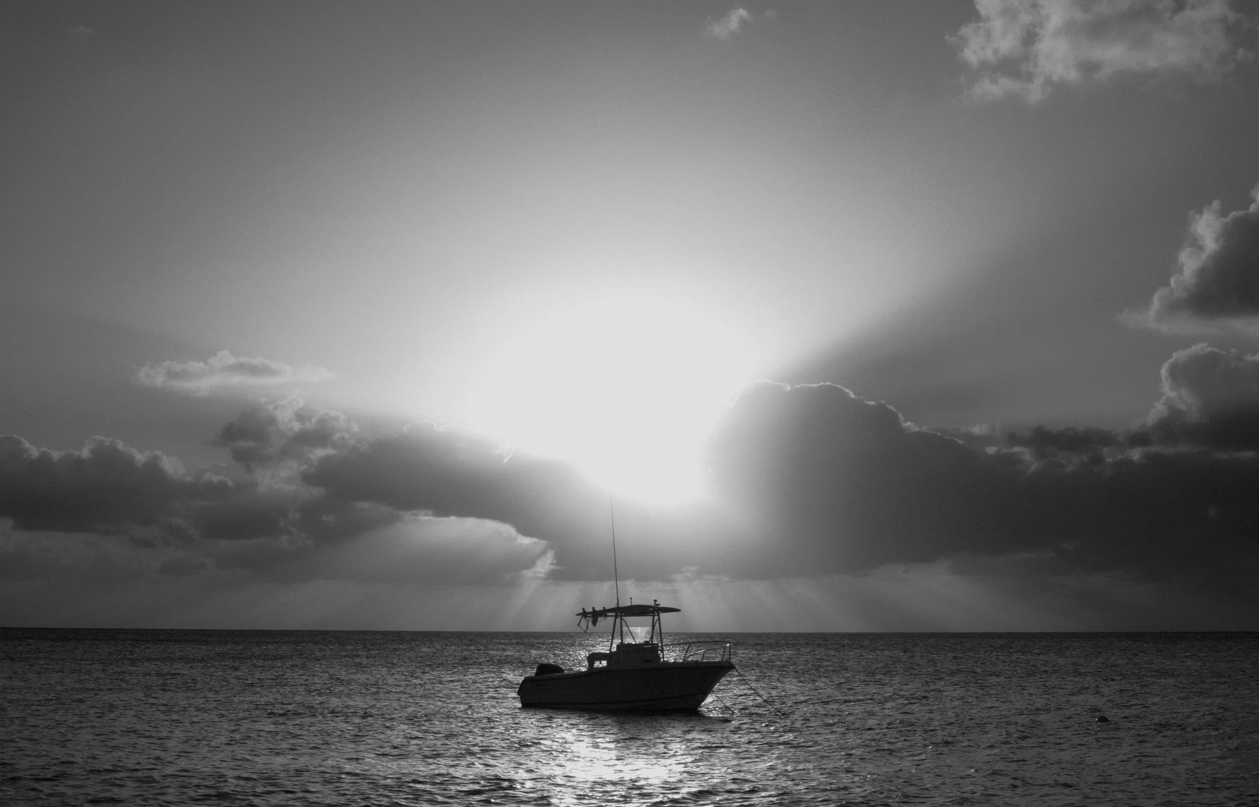 A black and white photo of a boat floating on the water with the sun setting behind clouds in the sky.