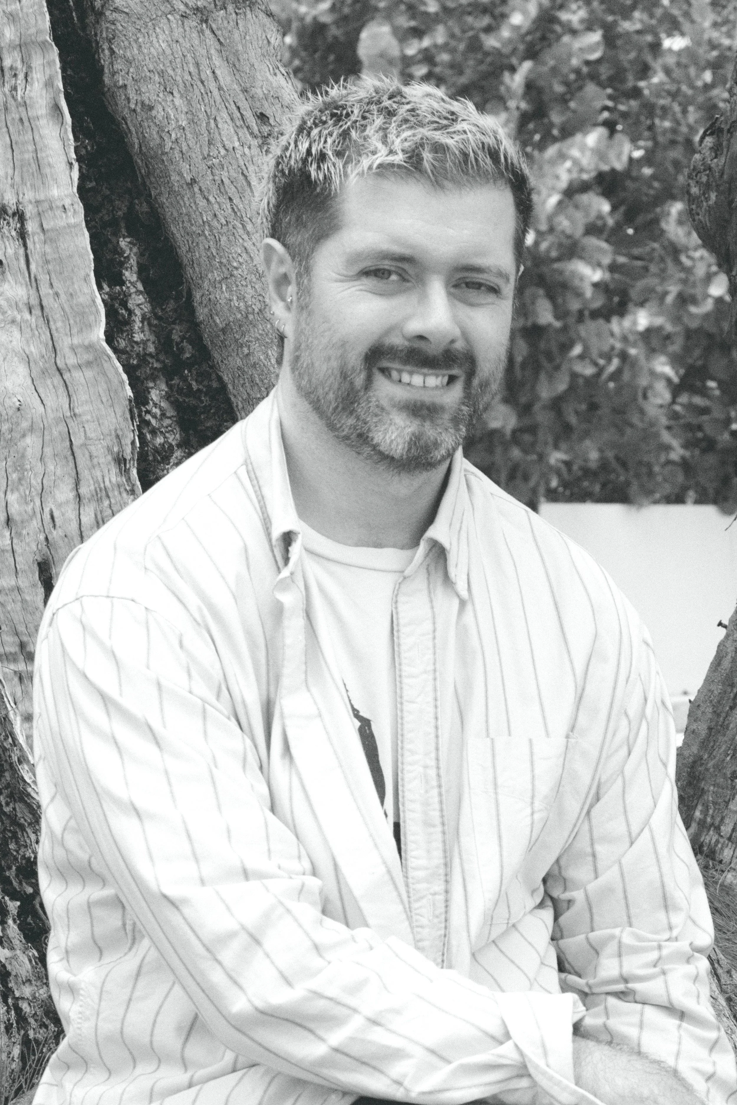 A man with short, light-colored hair and a beard, smiling at the camera, wearing a striped button-up shirt, sitting outdoors against the backdrop of trees and natural scenery.