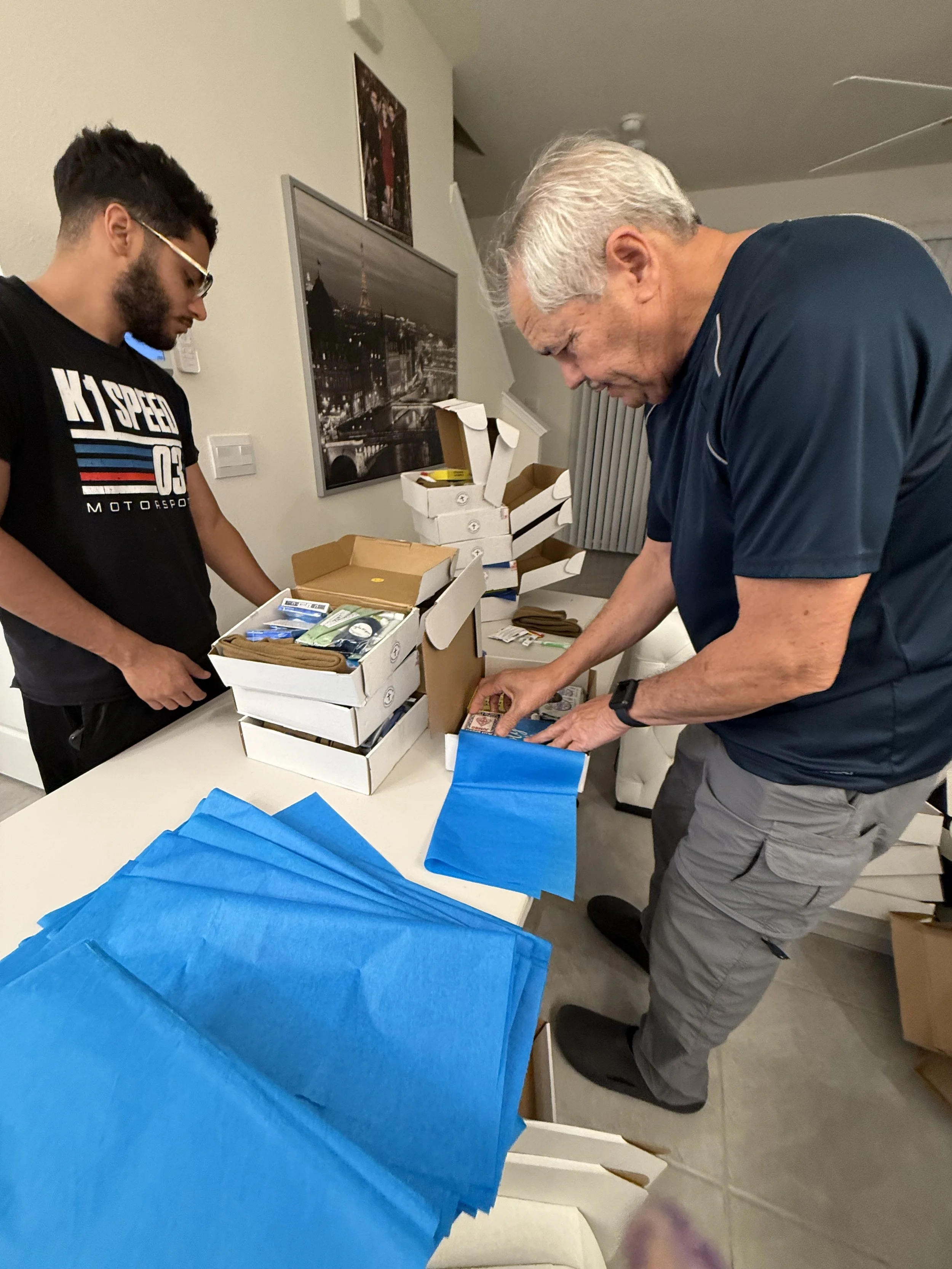Two men assembling packages at a table, surrounded by boxes, blue tissue paper, and various items.