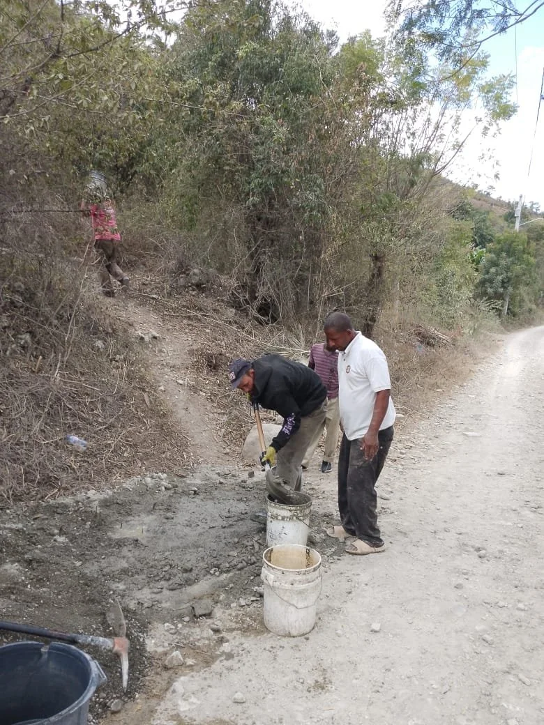 Two men working with cement and buckets on a dirt path next to trees.