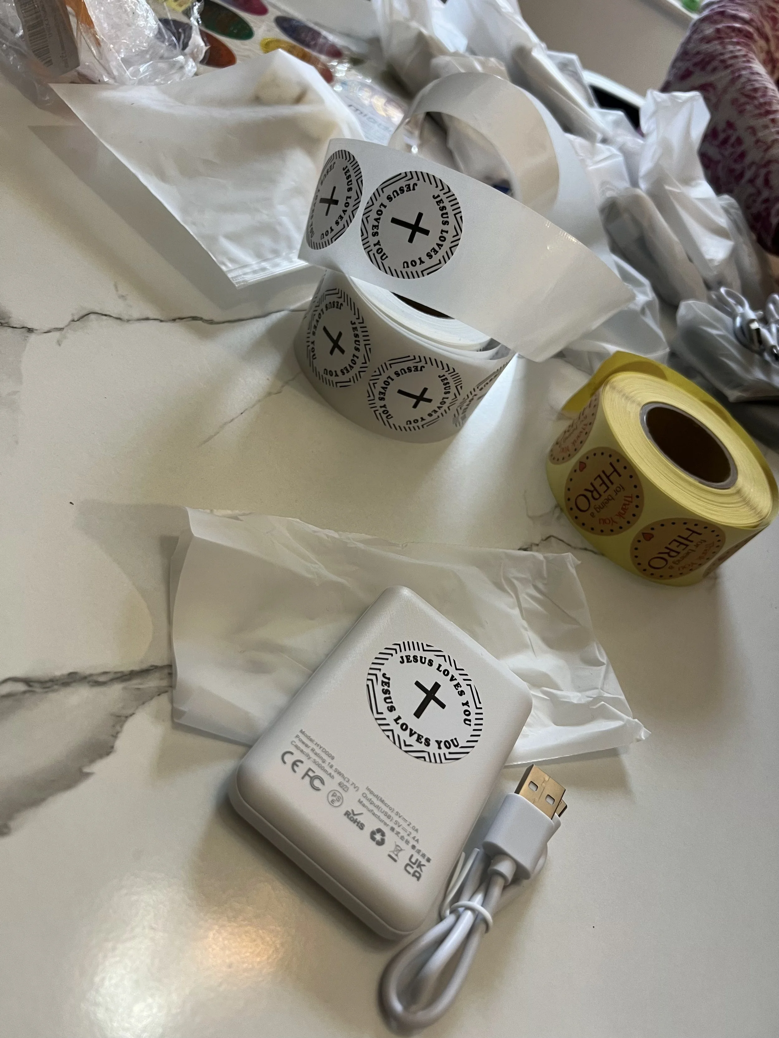 Rolls of stickers with "Jesus Loves You" text and cross design on a table, alongside a white power bank and USB cable.