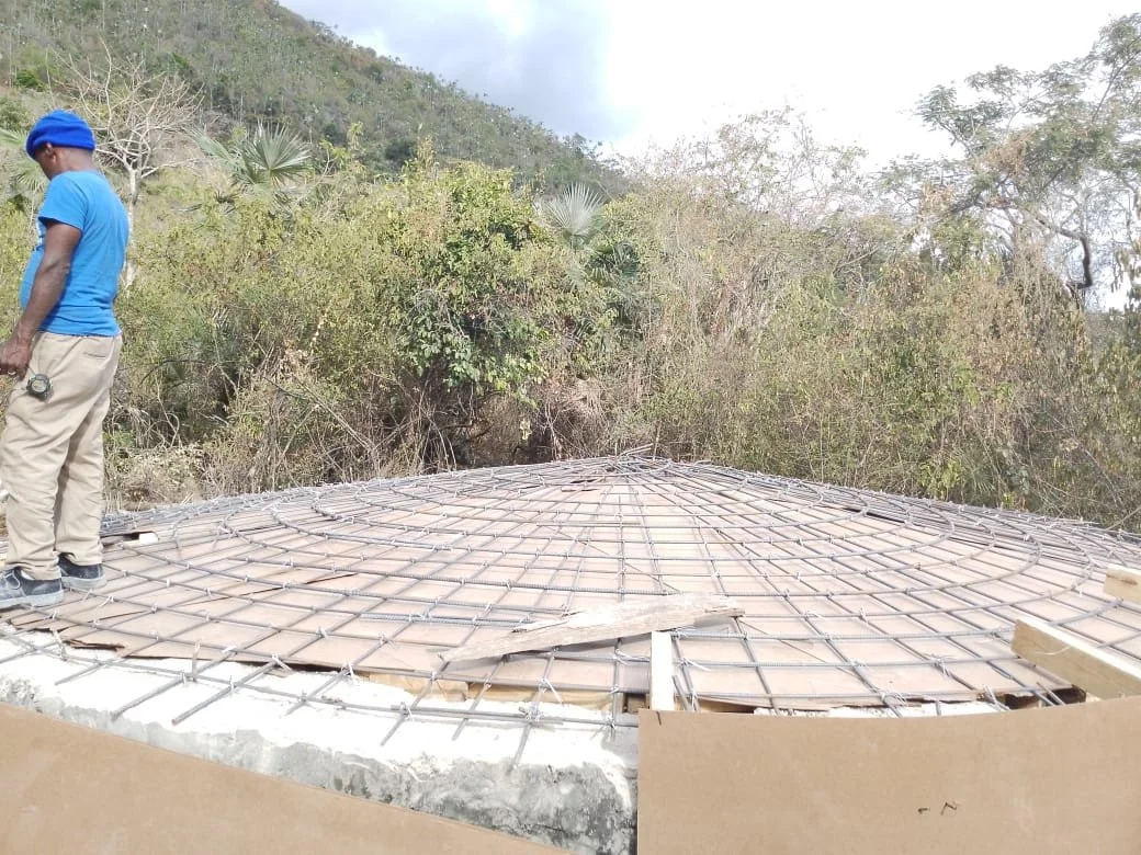Person standing on a circular concrete framework with rebar grid in an outdoor setting with trees and hills in background.