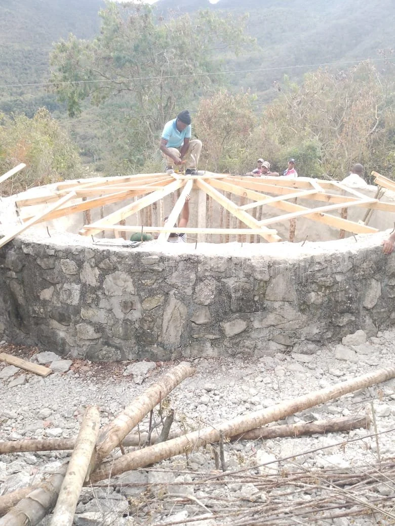 Construction worker building wooden roof frame on stone structure with trees and hills in the background.