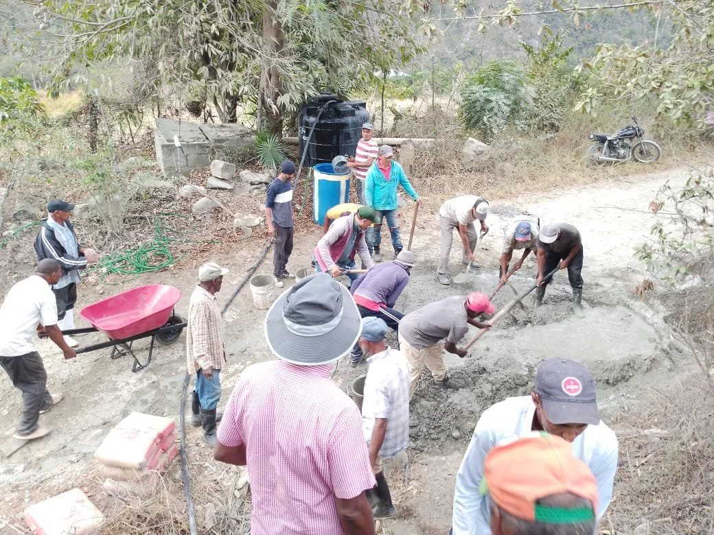 Group of people working on a water system, mixing and spreading concrete with tools in a rural outdoor setting, surrounded by trees and a motorcycle parked nearby.