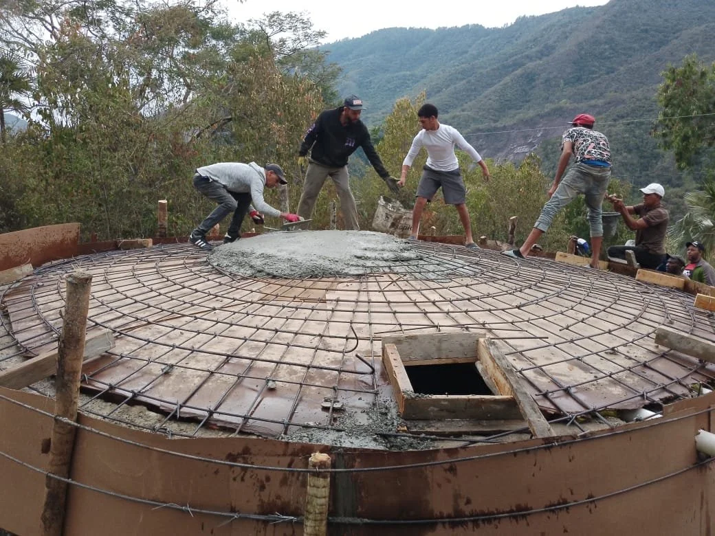 Construction workers pouring concrete on a dome structure with a rebar frame in a mountainous area.
